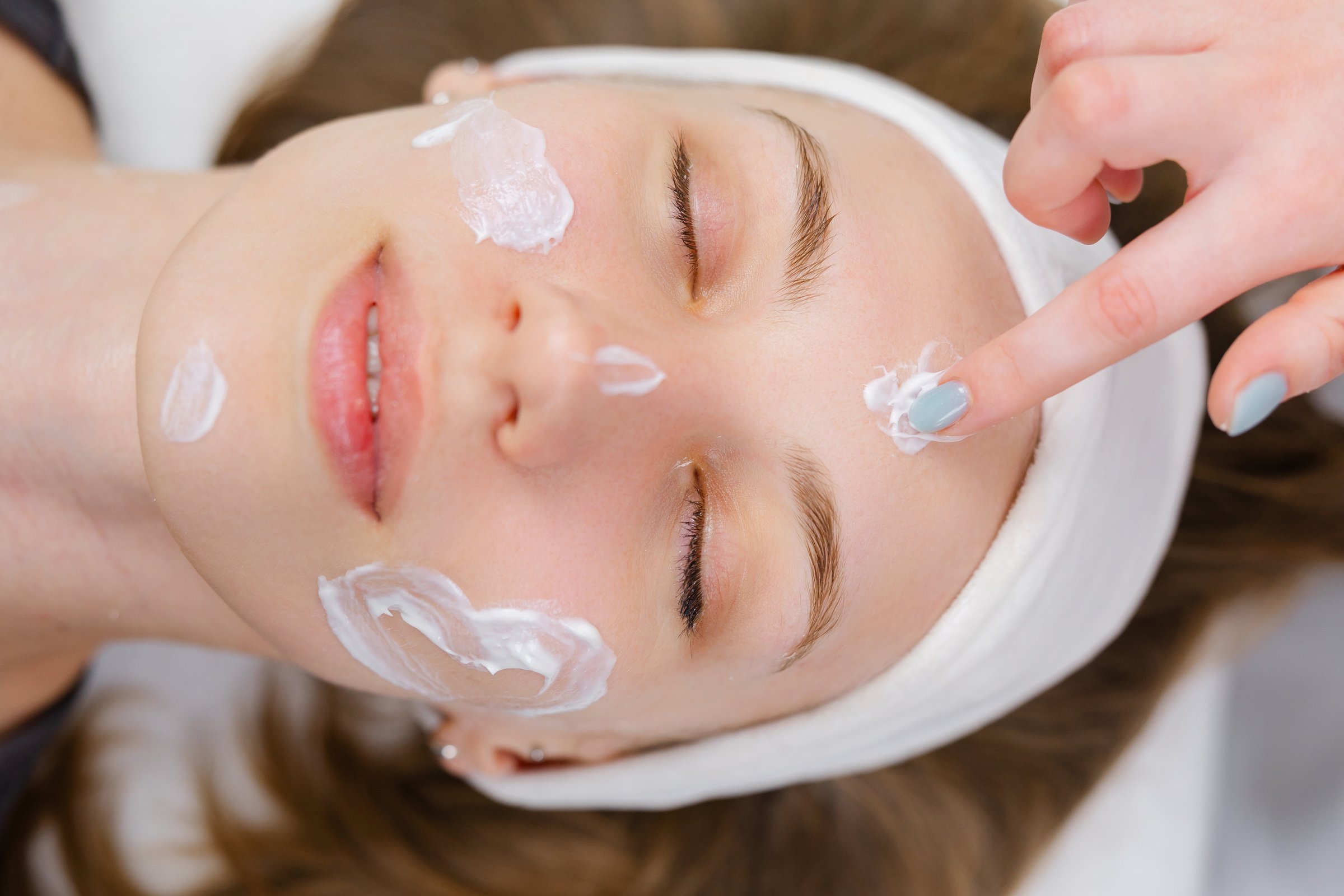 Beautician gently applying moisturizing cream on cheek of relaxed woman lying on a massage table with closed eyes during cosmetic face treatment in a beauty salon