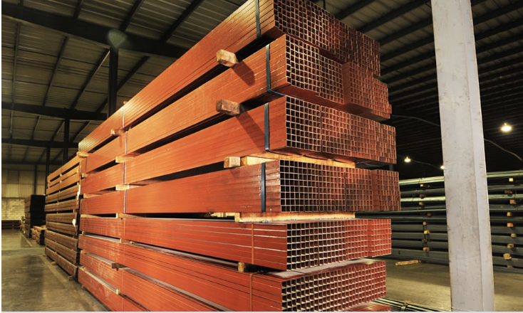 Stacked metal square tubes painted red in an industrial warehouse setting with lighting overhead.