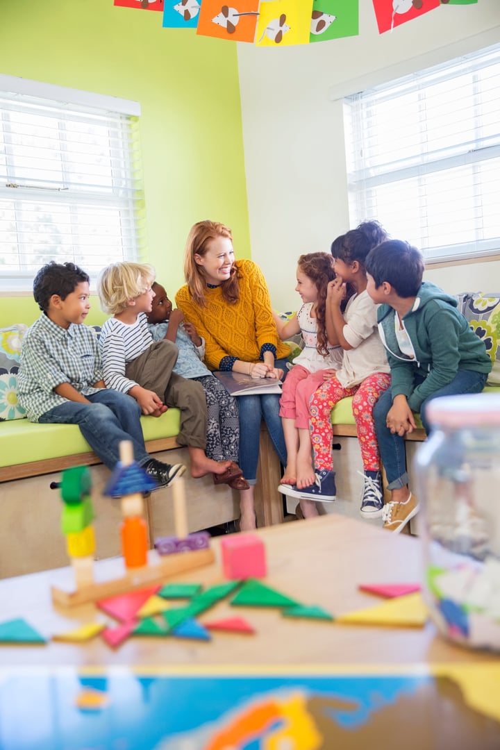 Teacher sitting with group of diverse children on bench in a brightly colored classroom, engaging in conversation and play.