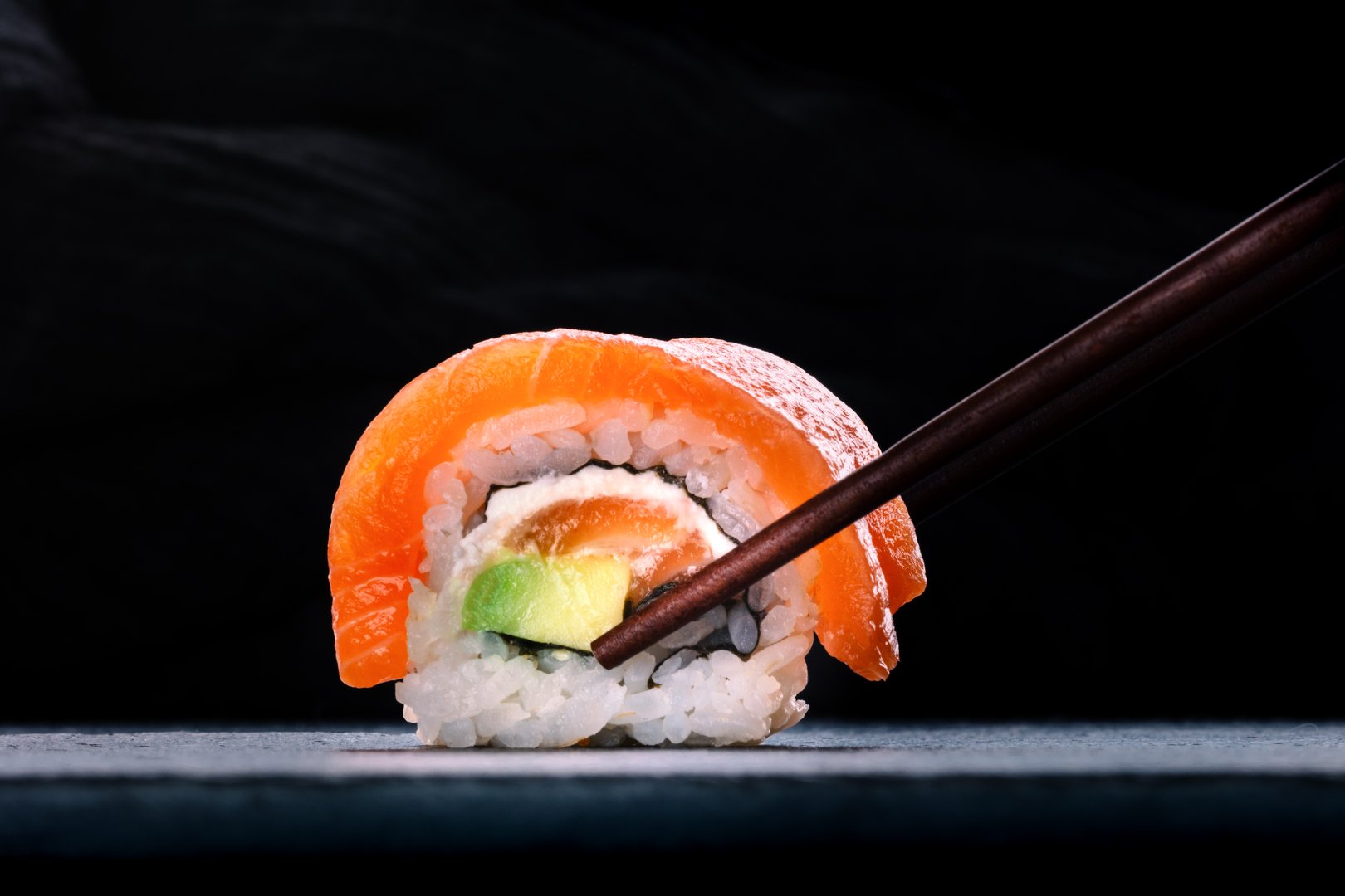 Salmon sushi with avocado and cream cheese, uramaki roll on a black slate background, with chopsticks