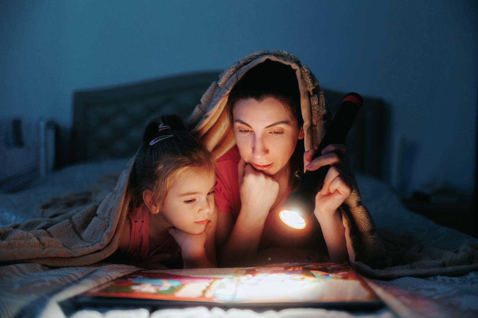 Sharing special moments, a mother and her little girl enjoy reading a bedtime story together by flashlight, wrapped in warmth under the covers at night.