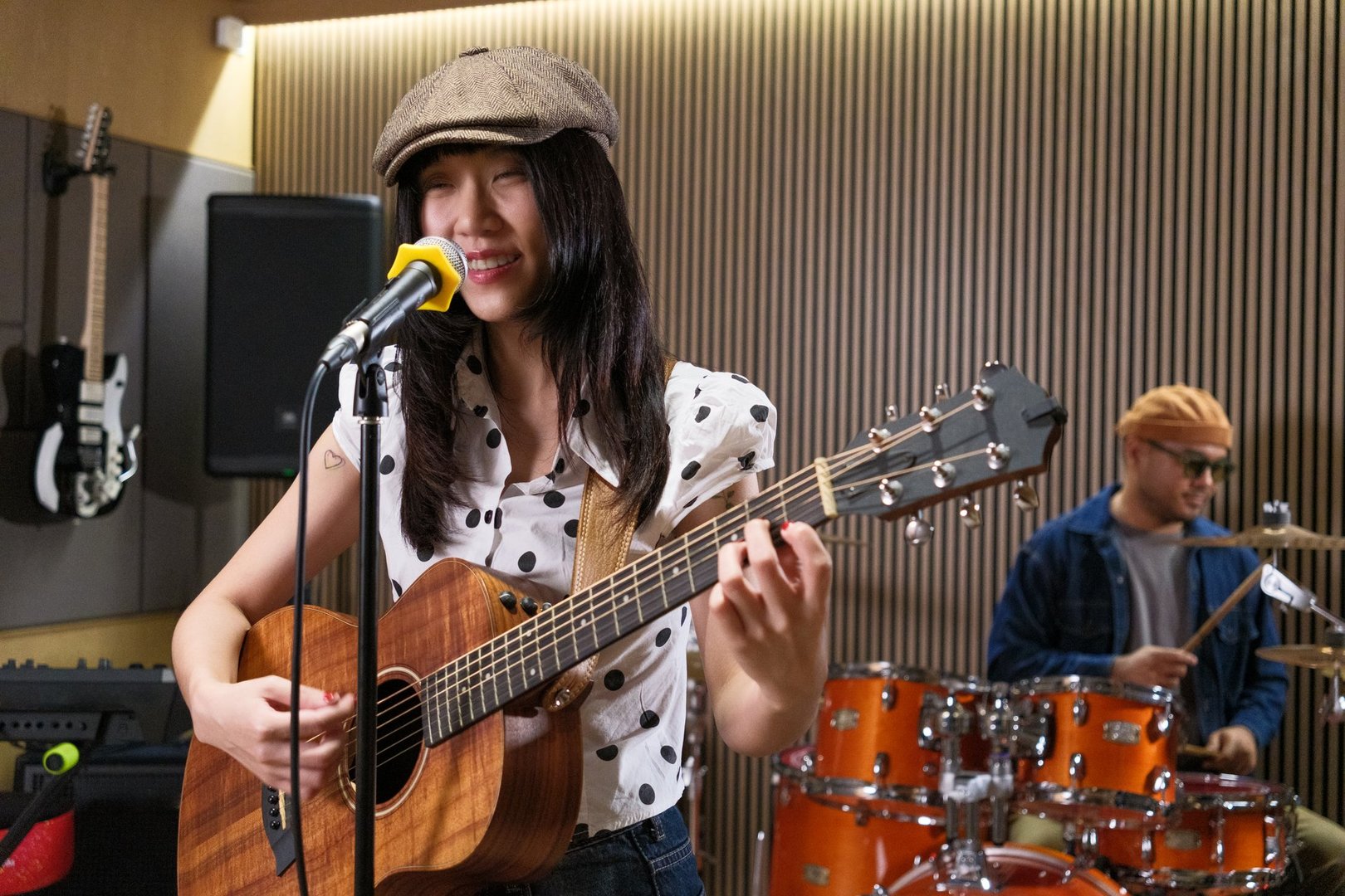 Professional asian female musician is happy singing and playing acoustic guitar while a male drummer play in the background.