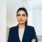 Person in a business suit stands confidently against a light-colored wall in a well-lit room.