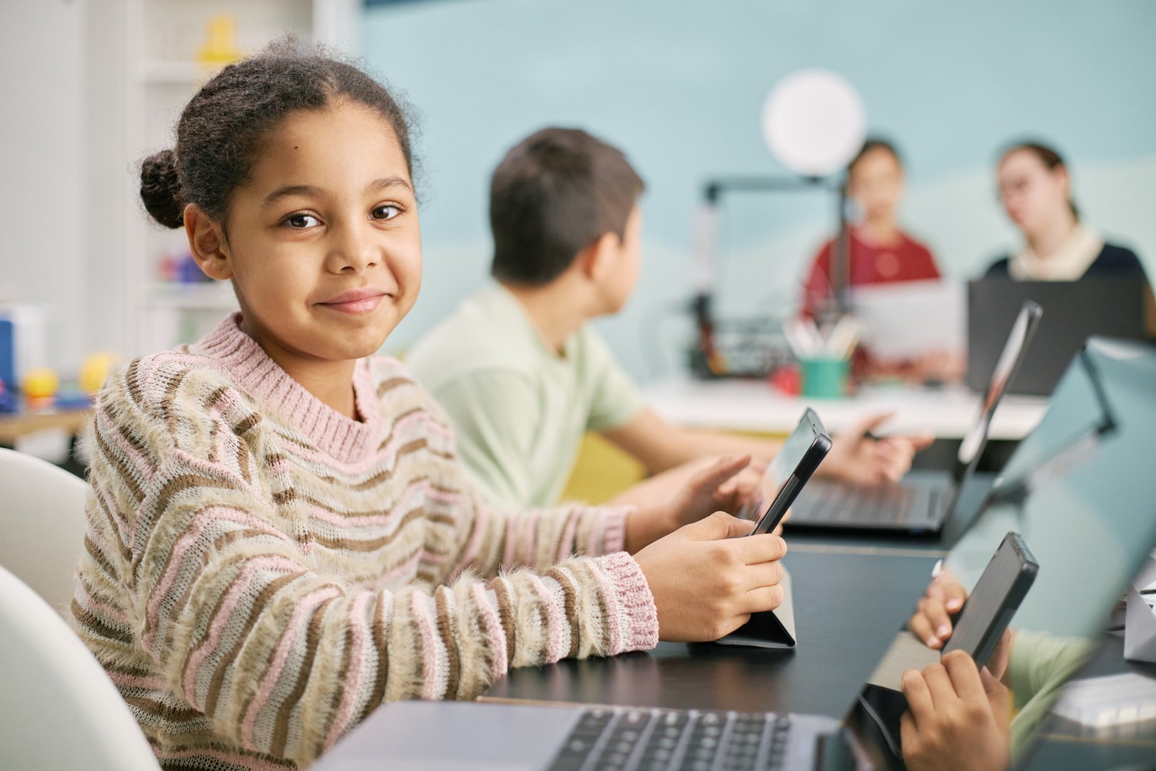 Group of multi-ethnic children seated around a table, engaging with laptops and tablets in an educational setting. Smiling child in focus, looking directly at camera