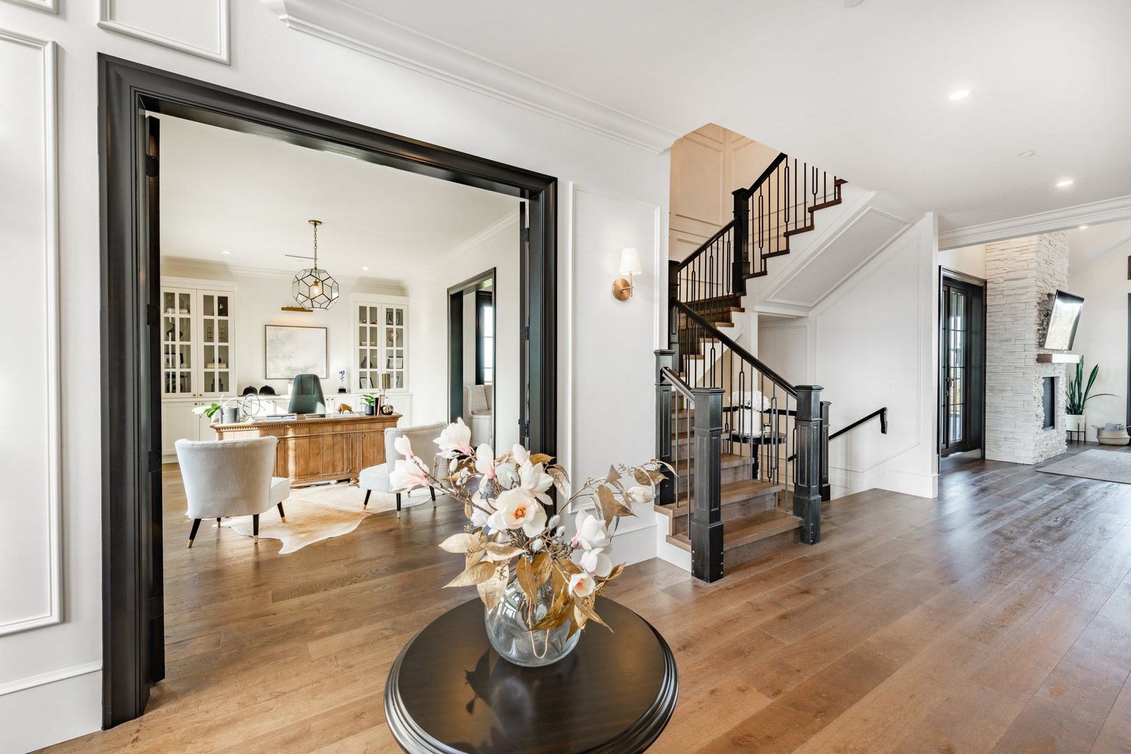 Beautiful large and luxurious estate home interior front door entry hallway foyer with large glass and black wood door expansive entry with a round table containing an arrangement of dried flowers in a bouquet. The view from the front is to a home office and a casual sitting room with blue velvet sofas. large wooden barn doors lead to another room there is a staircase and steps going up.