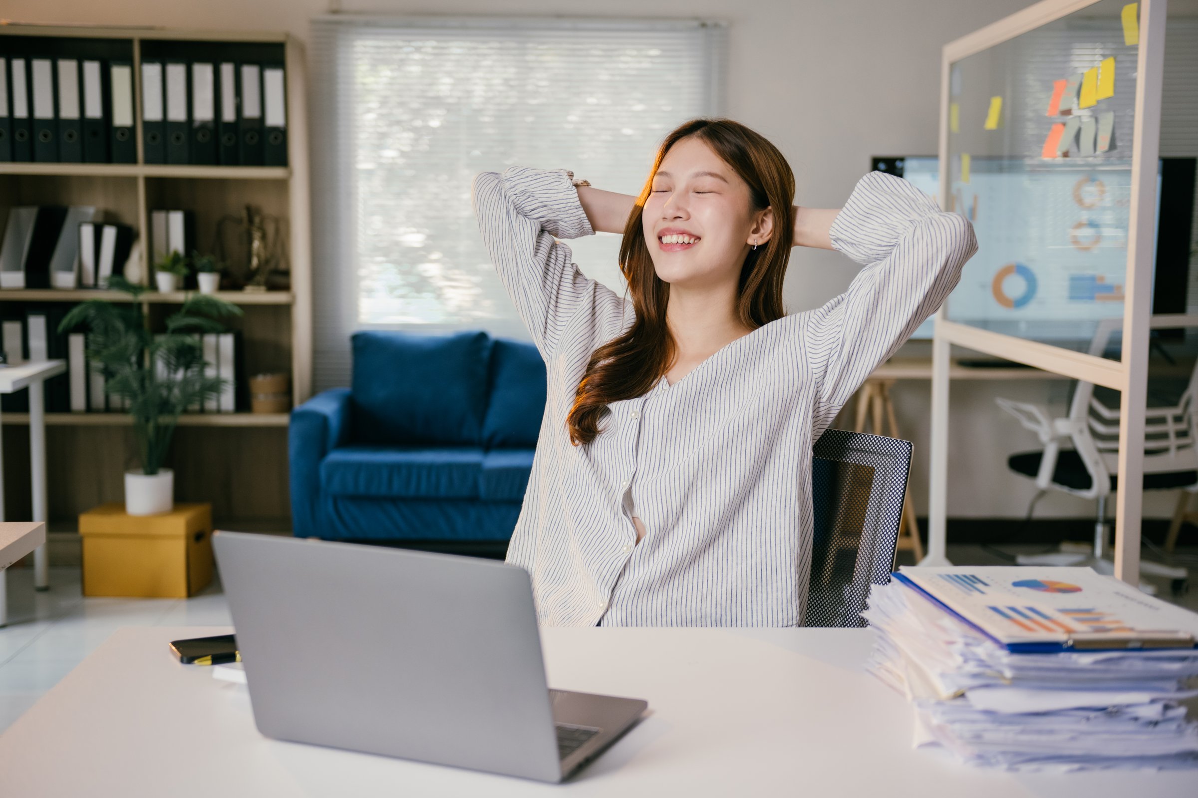 Young businesswoman is sitting at her desk with her hands behind her head, relaxing after finishing work on her laptop, with a pile of paperwork next to her