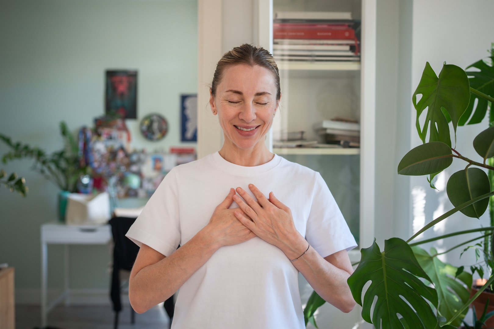 Happy peaceful middle aged woman standing alone indoor with closed eyes keep hands close to heart with hope pray in mind. Emotional female smiling feeling pleasure in deep gratitude. Thank you life.