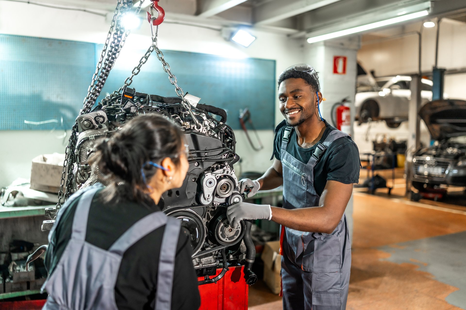 Two smiling mechanics are working on a car engine suspended by a chain hoist in a professional garage