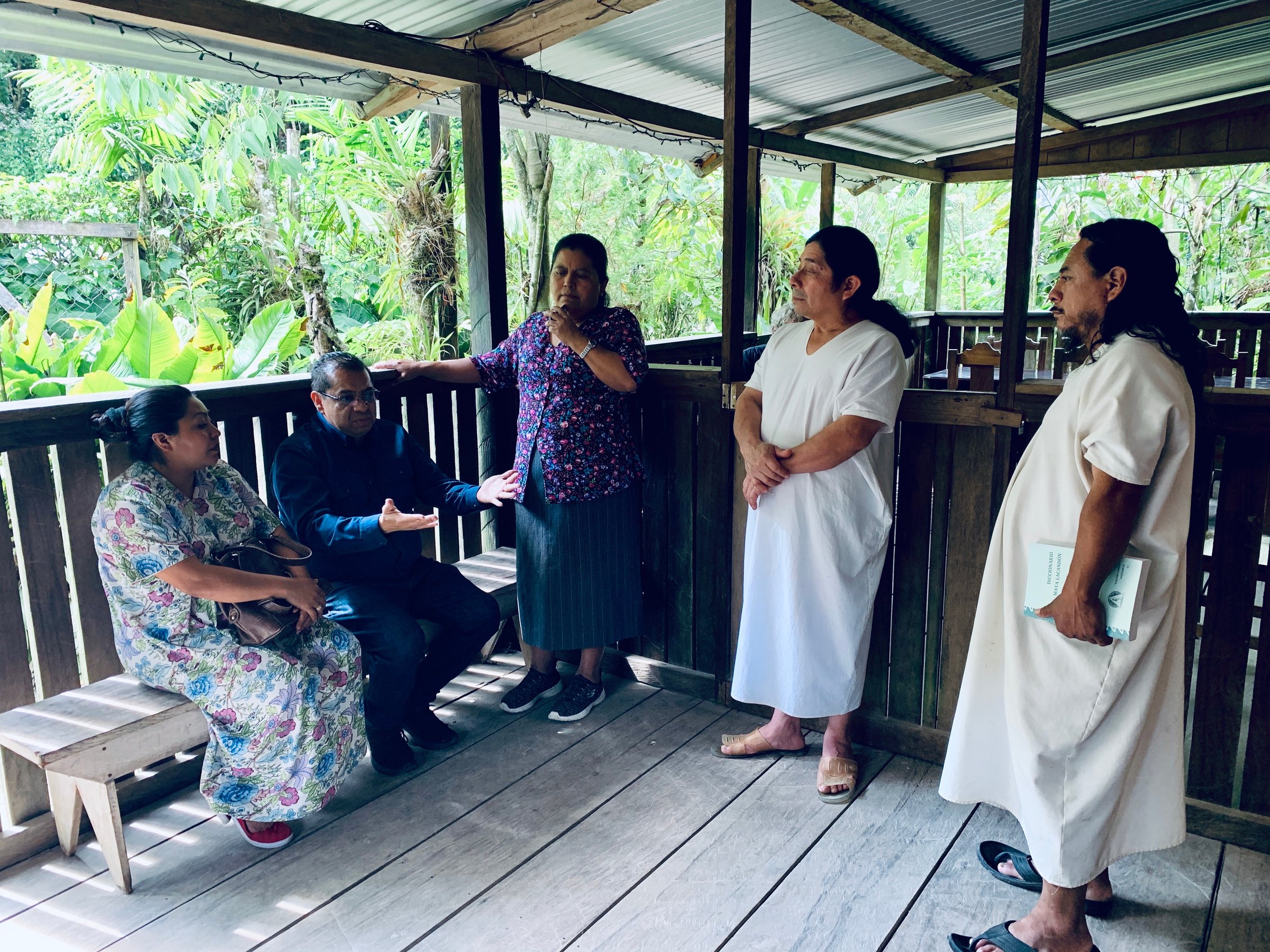 A group of people engaged in conversation on a wooden deck surrounded by lush greenery.