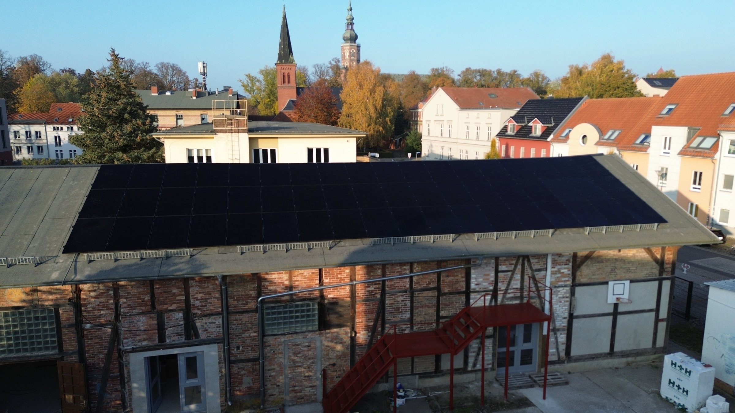 Rooftop solar panels on an industrial building with autumn trees and church steeples in the background.