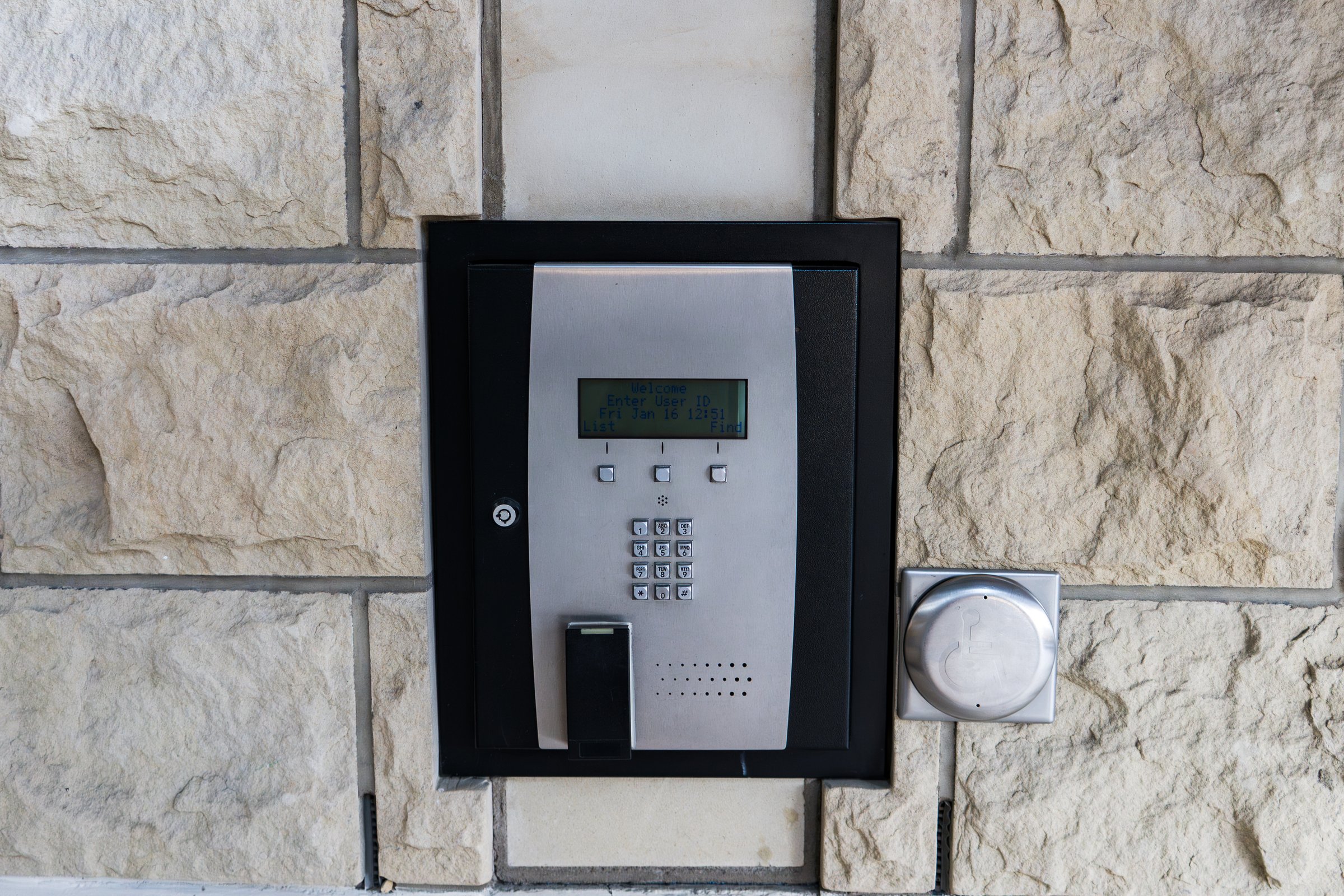 A modern security system seamlessly integrated into a textured stone wall. The panel includes a keypad, display, and intercom, providing access control to the building.