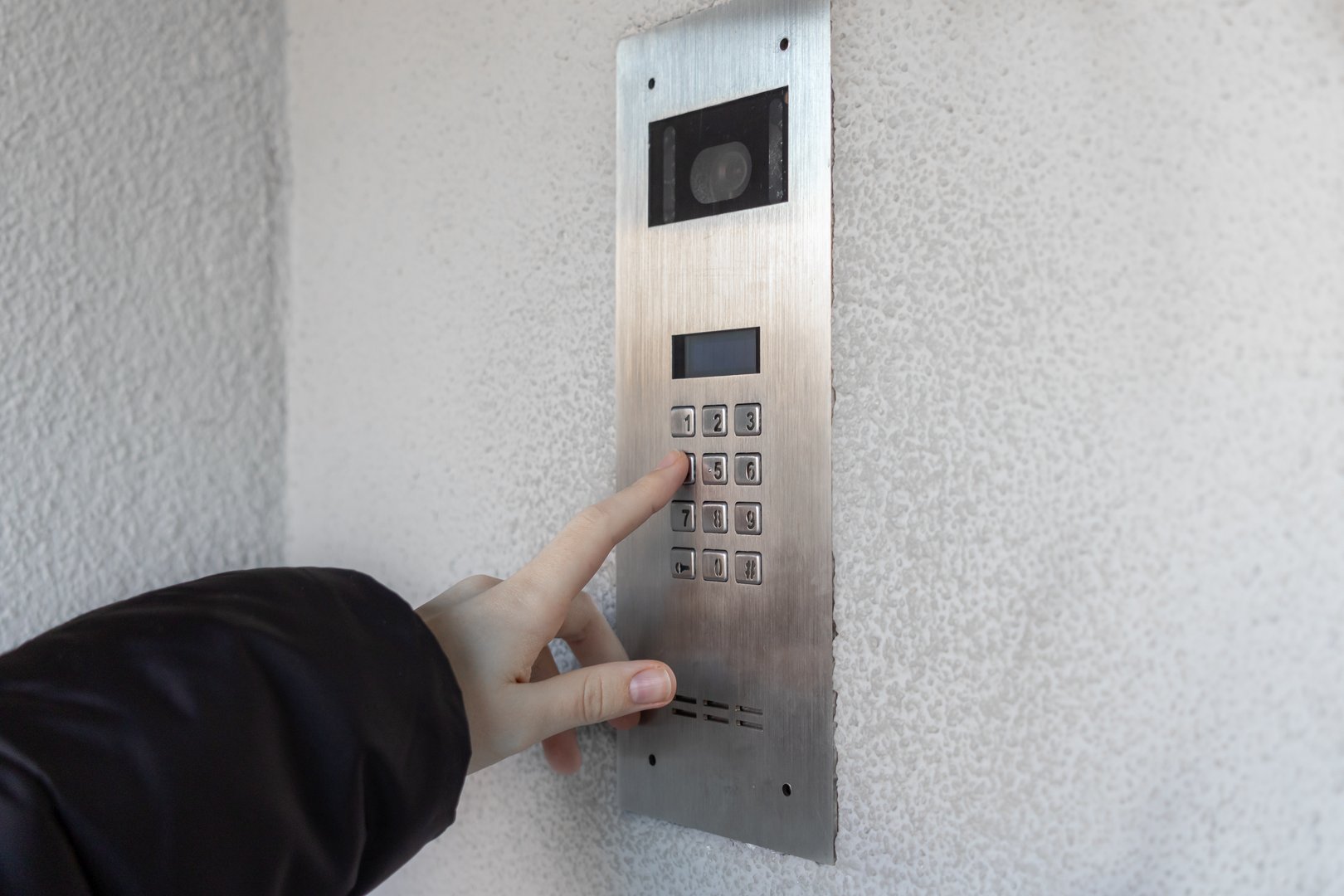 Hand pressing button on modern intercom keypad with camera on textured white wall for secure building access