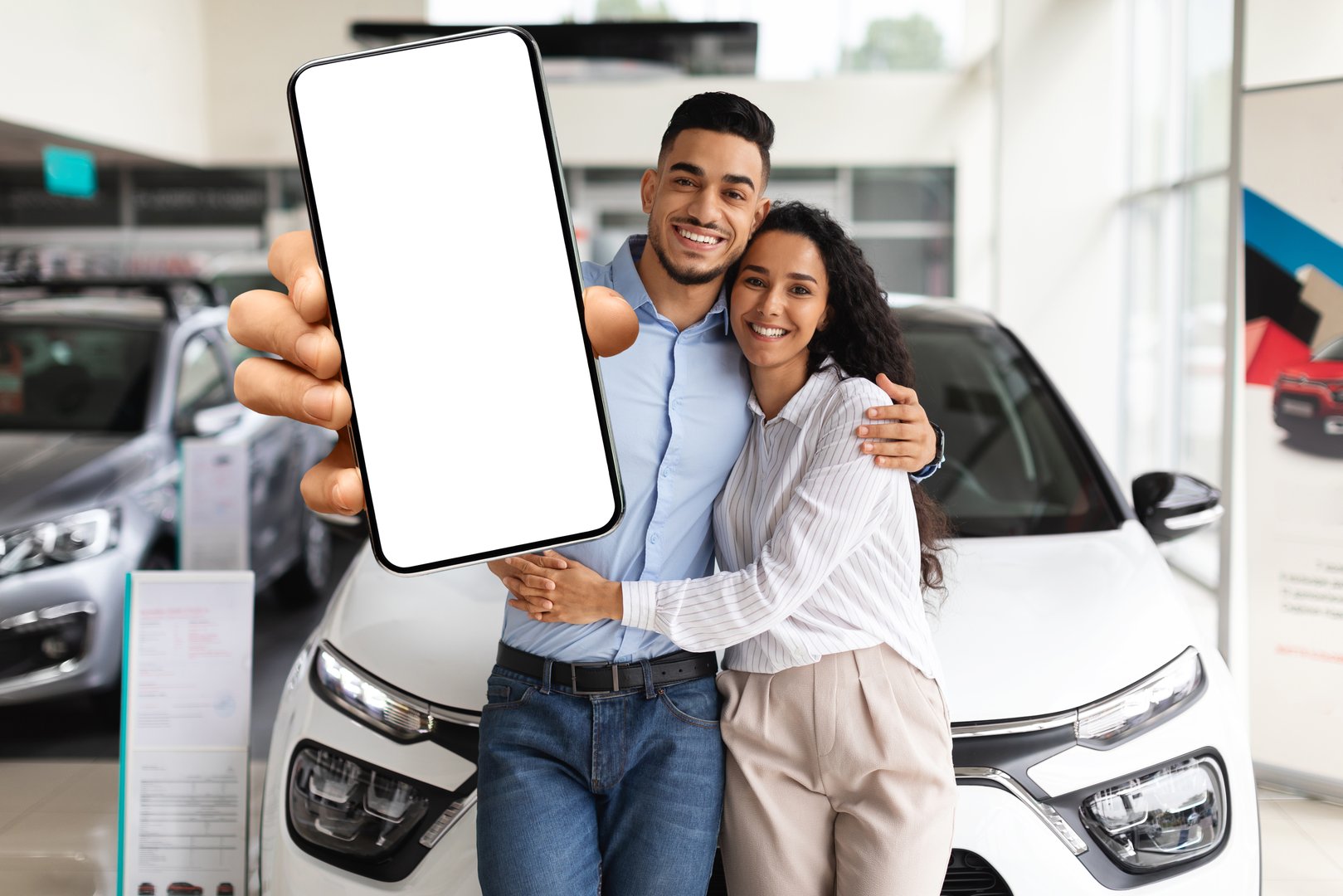 Happy middle eastern couple young man and woman standing by nice brand new car, showing smartphone with blank screen, embracing and smiling, buying new auto together at luxury showroom, mockup