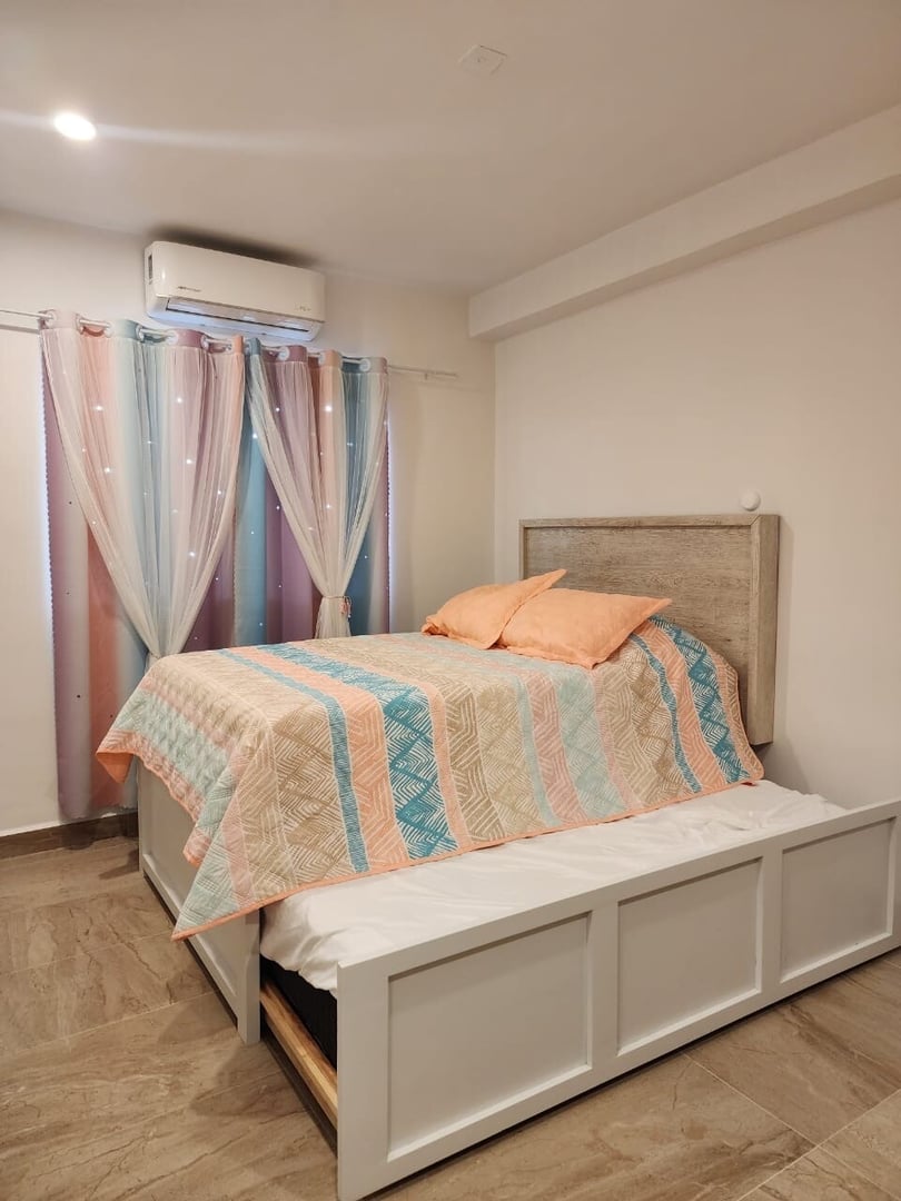 Bedroom with a double bed, pastel-striped bedding, sheer curtains, tiled floor, and air conditioning unit above the window.