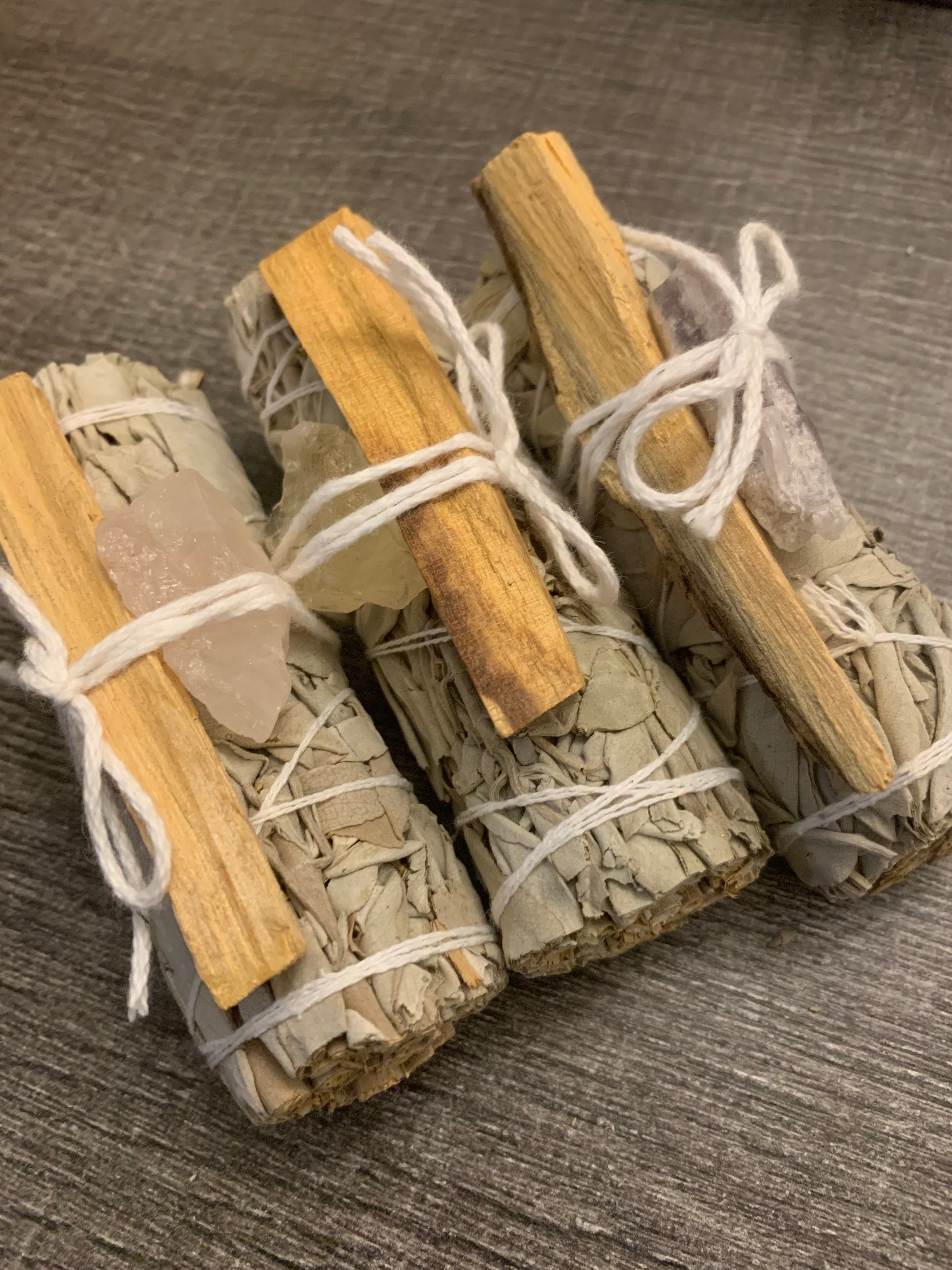 Bundles of sage wrapped with white string, each with a palo santo stick and a crystal, on a textured surface.