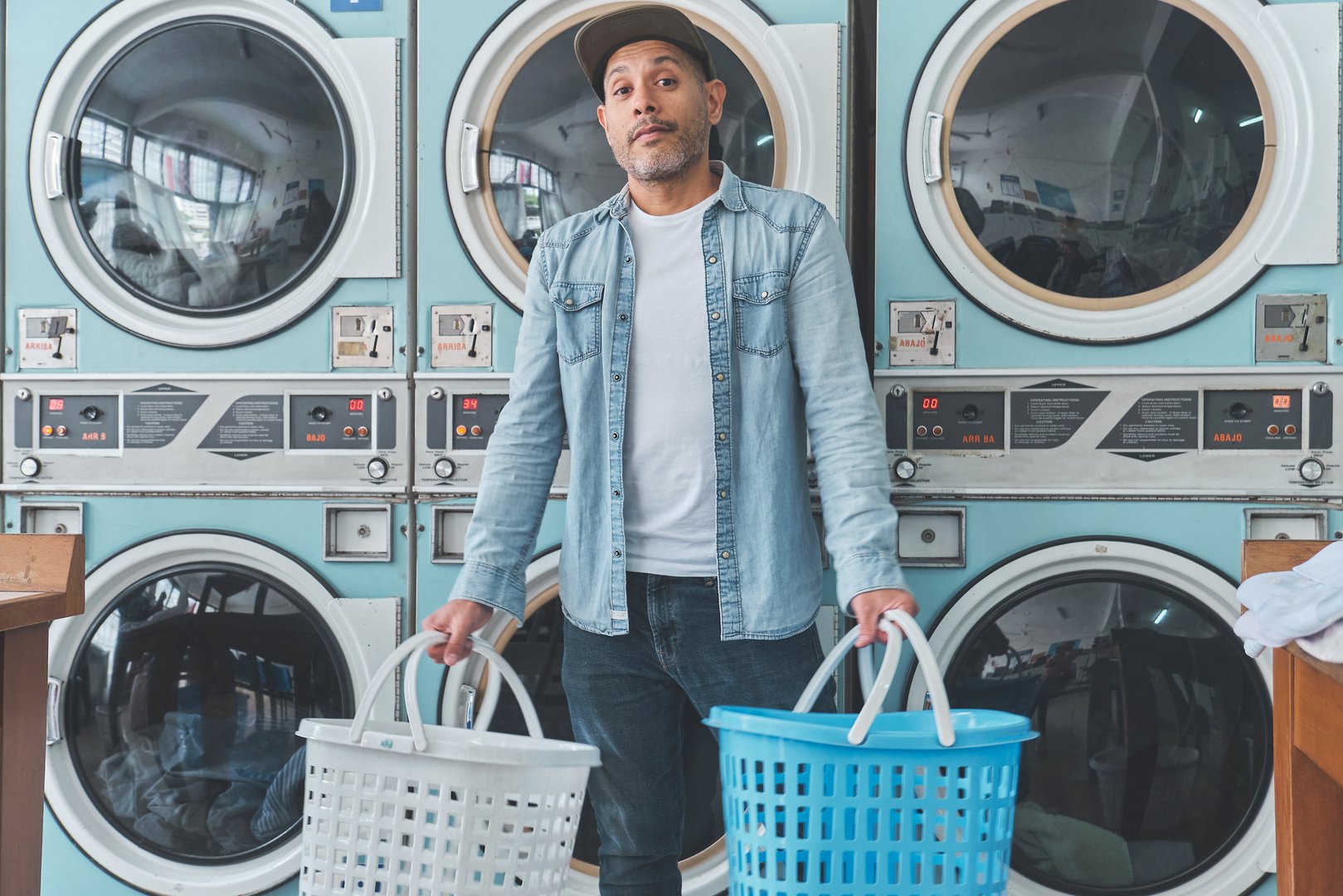 Young man is holding two laundry baskets, smiling and looking away, in a laundromat full of washing machines