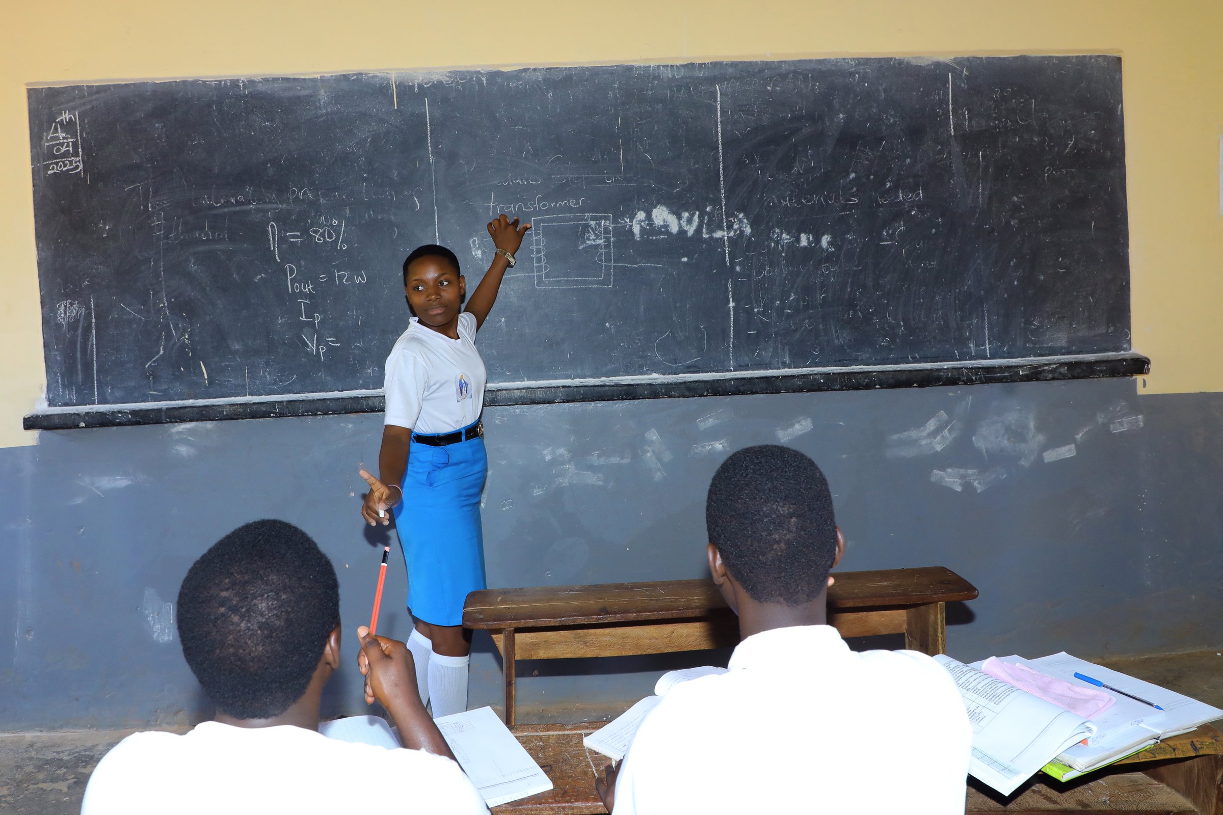 A student in uniform explains a topic on a chalkboard while two classmates take notes in a classroom.