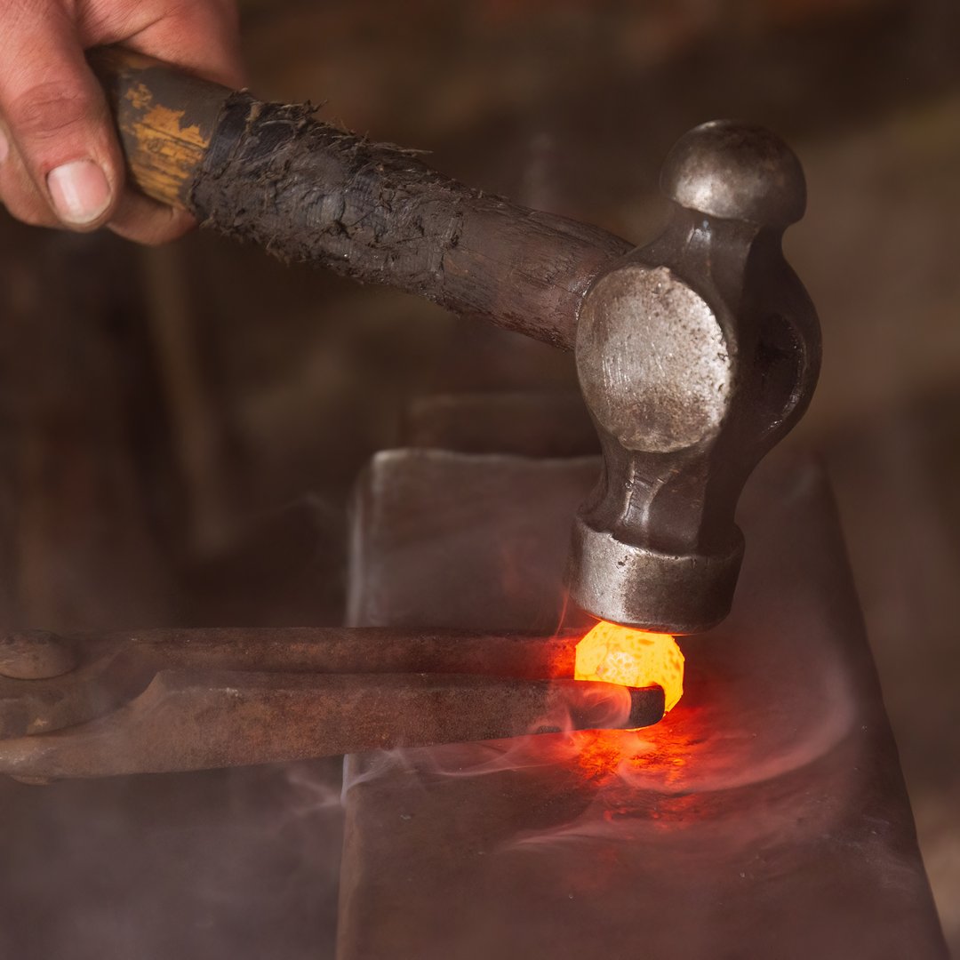 A hammer being used to shape a sphere of red hot steel held by tongs on an anvil in a forge