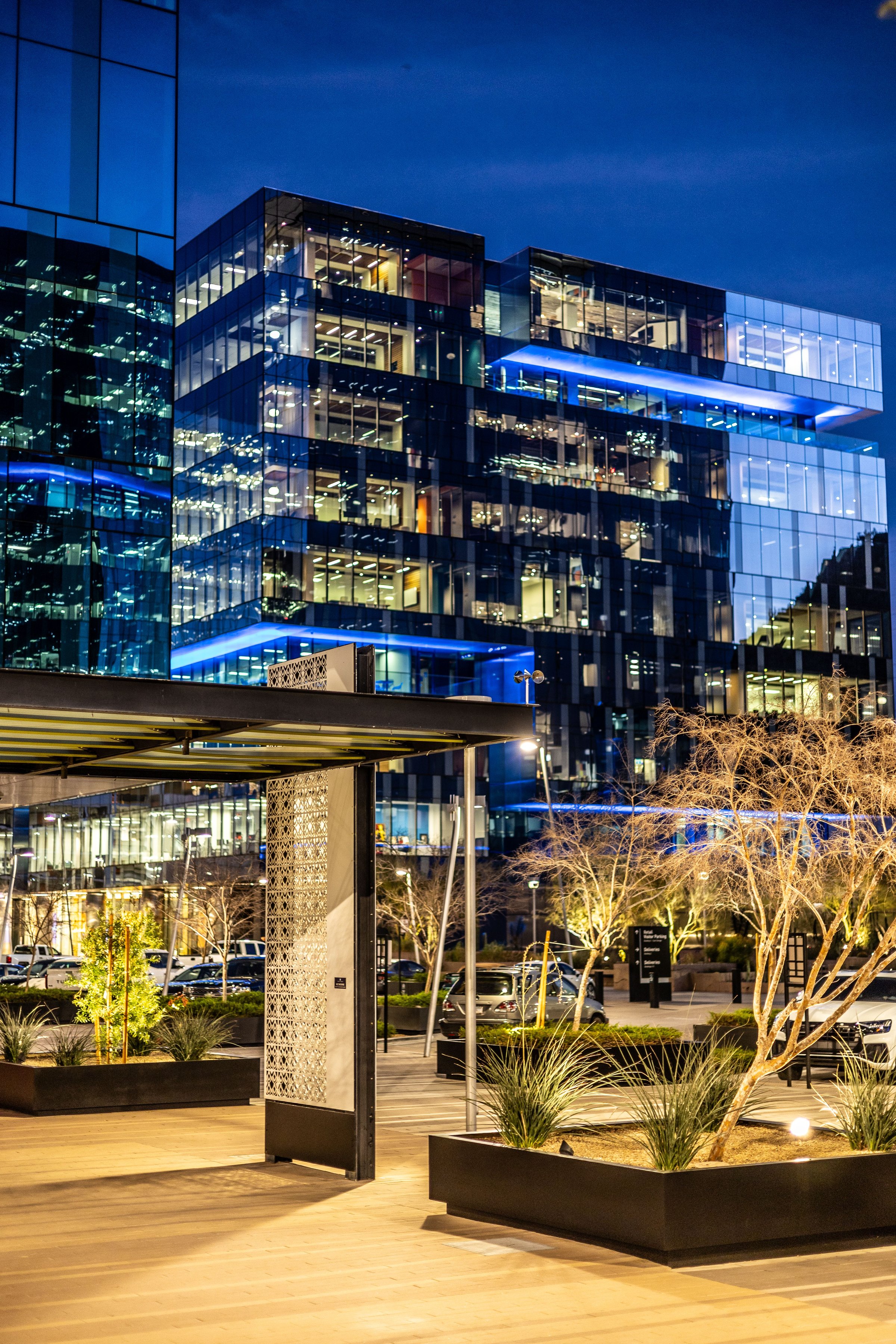 Modern office building with illuminated glass facade at night, surrounded by landscaped urban area.
