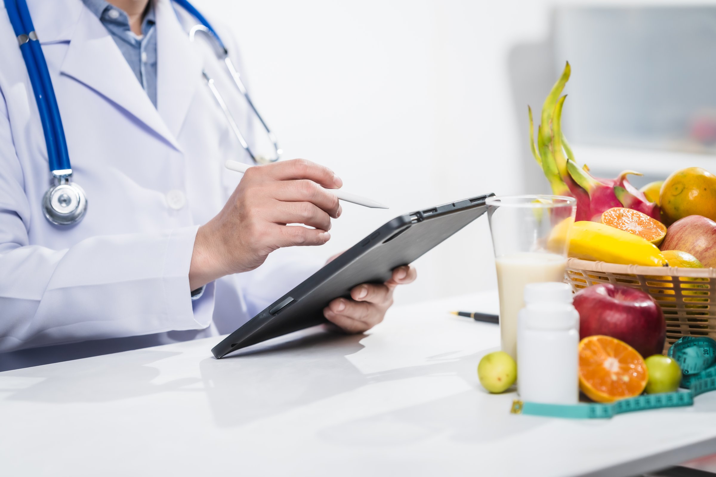 A male nutritionist working at a hospital desk, holding an apple and mixed fruit, offering care, health advice, weight loss guidance, and eating tips online for patients seeking nutrition support