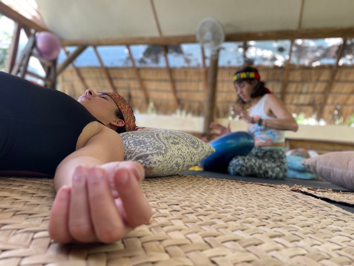 Person lying on a mat, eyes closed, while another person plays a musical instrument in a bamboo structure.