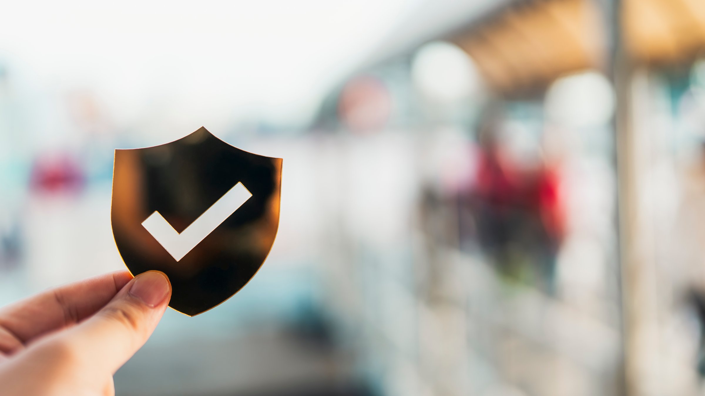 Hand holding shield with check mark icon in front of blurred people walking background, symbolizing data protection, security, safety assurance, travel safety, and digital privacy concept.