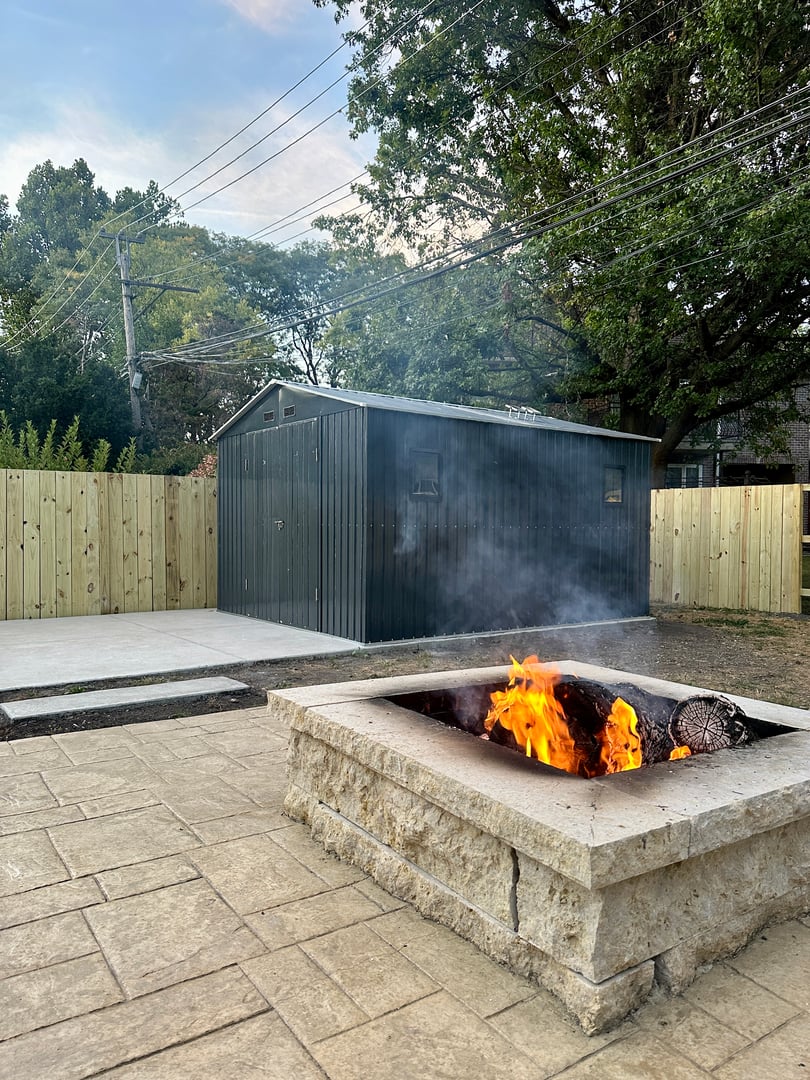 Outdoor fire pit with bright flames, near a green shed and fenced yard, surrounded by trees and utility lines in the background.