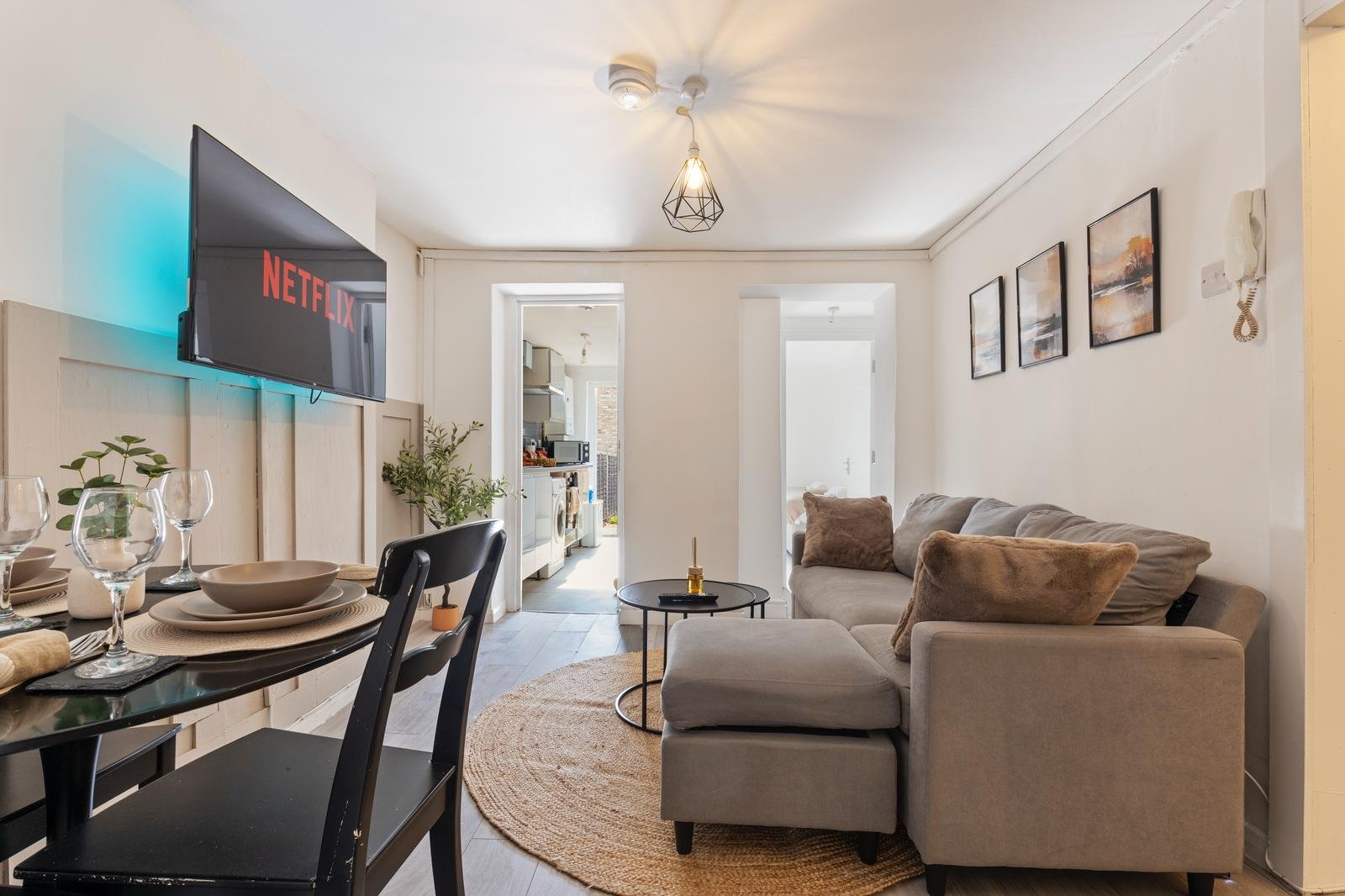Modern living room with gray sectional sofa, wall-mounted TV, and dining table. Light decor and wall art enhance brightness.