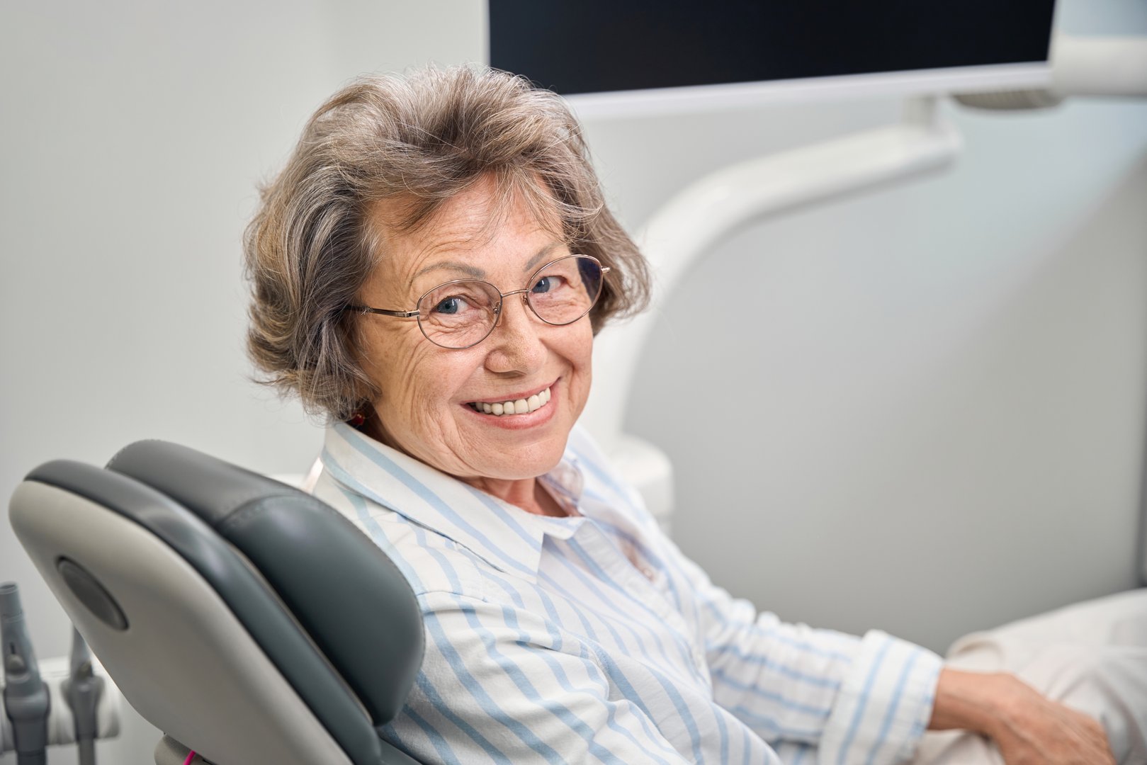 Gray-haired elderly woman sits in a dentists chair, lady in glasses