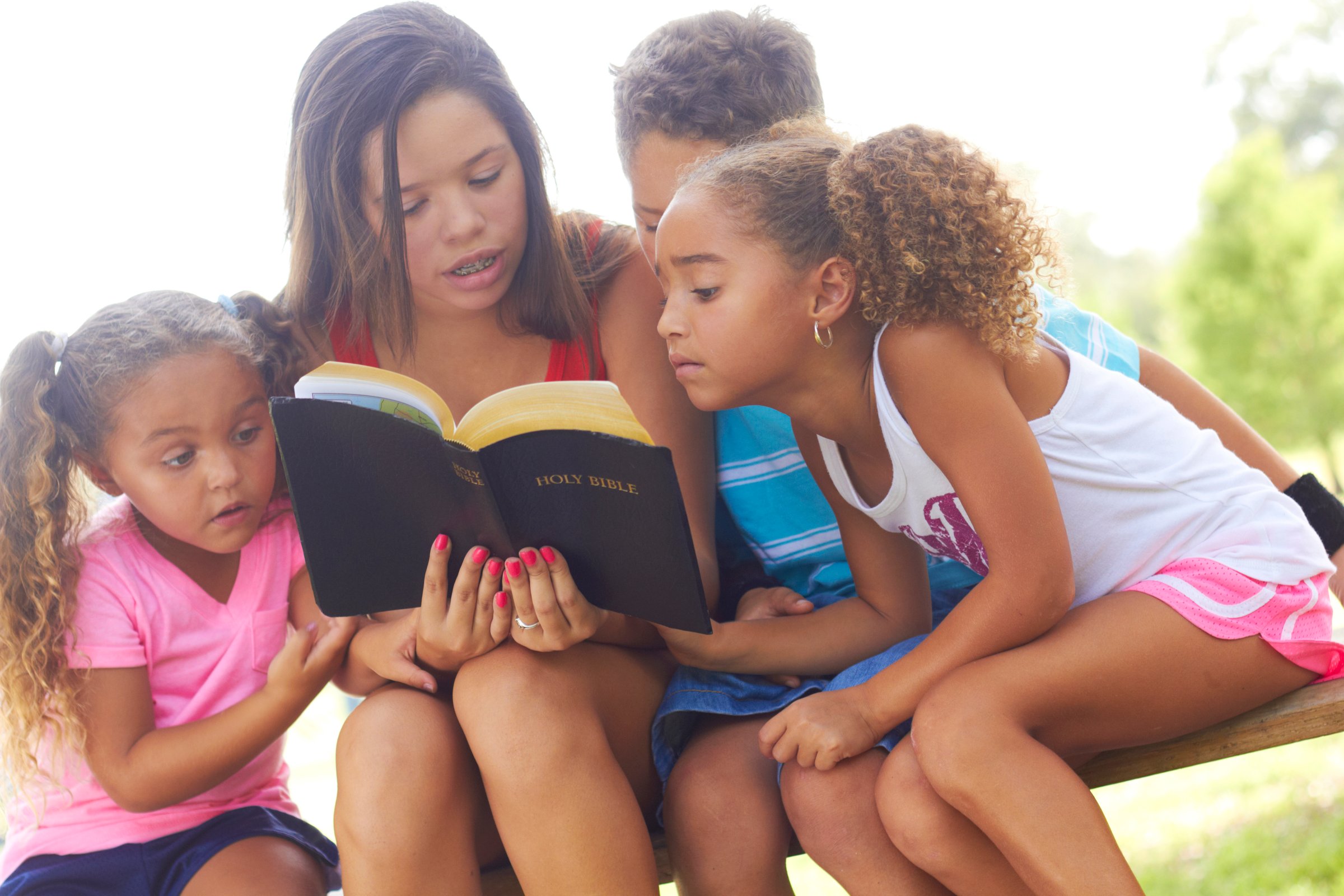 Teenage girl reads to her three siblings from the Holy Bible on park bench. Horizontal shot.