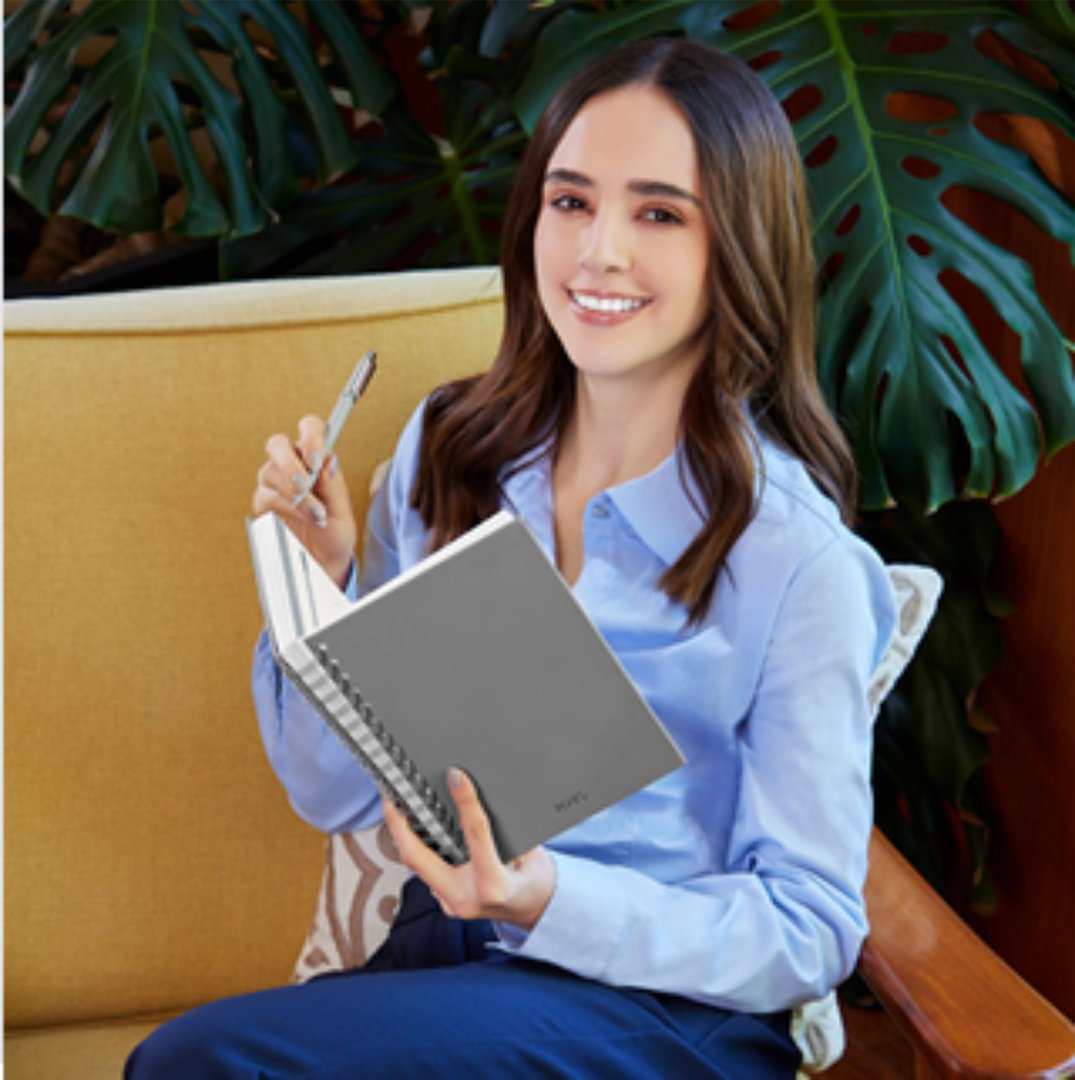 Woman with long hair smiling, holding a pen and open notebook, sitting on a yellow couch with a tropical plant behind her.