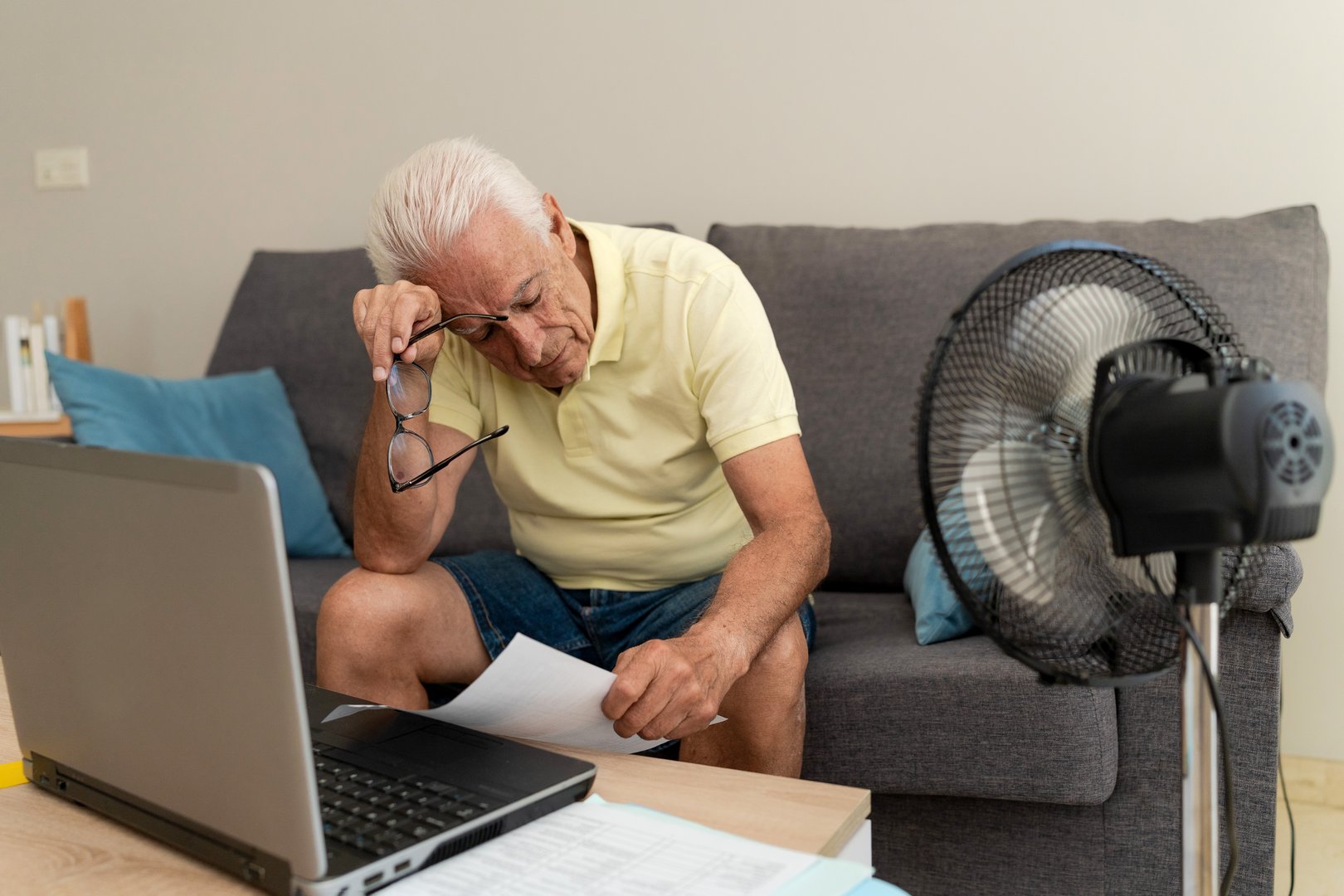 Worried senior man calculating domestic bills at home, using laptop and cooling off with a fan