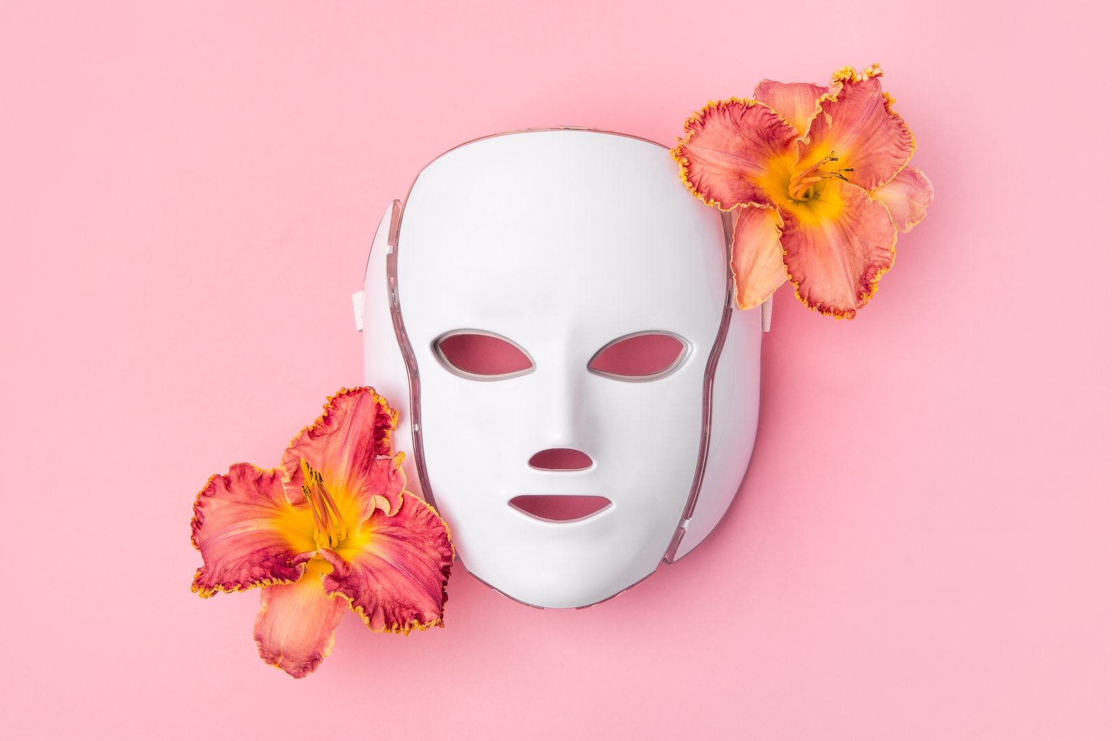 Cosmetic LED facial mask regenerative treatment on pink background with flowers lilies. Health and beauty of the face.