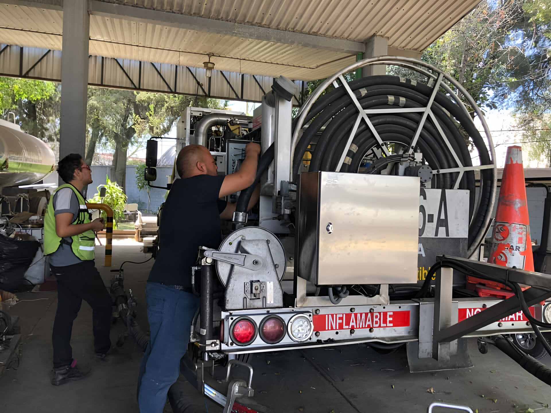 Two workers inspect and adjust equipment on a large cable reel truck in an industrial outdoor setting.