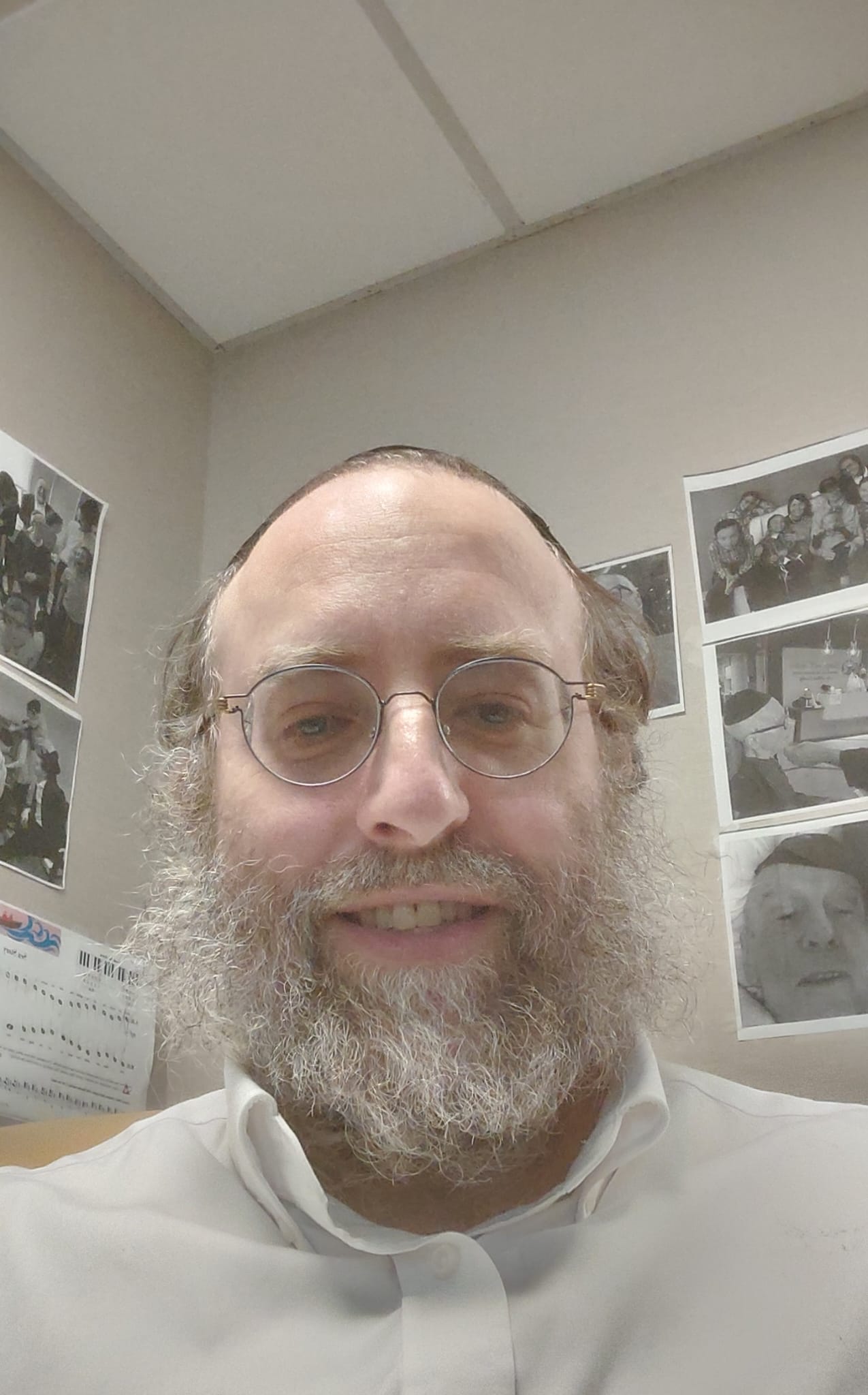 A bearded man with glasses smiling, sitting in an office with photos on the wall behind him.