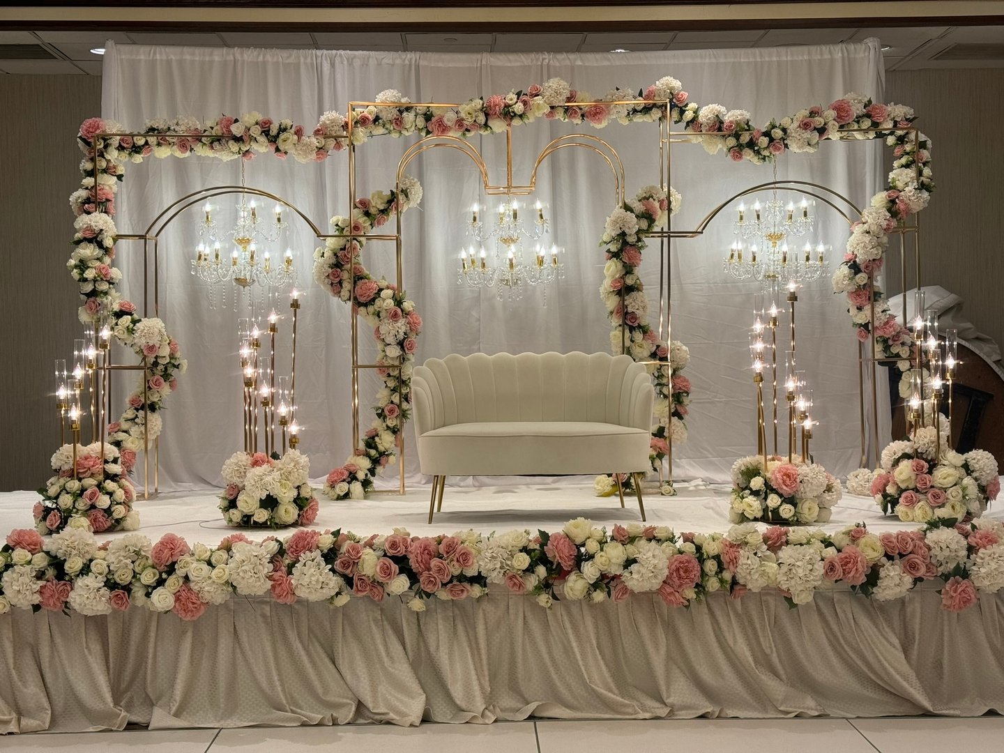 Elegant wedding stage with floral arch, white couch, and chandeliers, adorned with pink and white flowers.