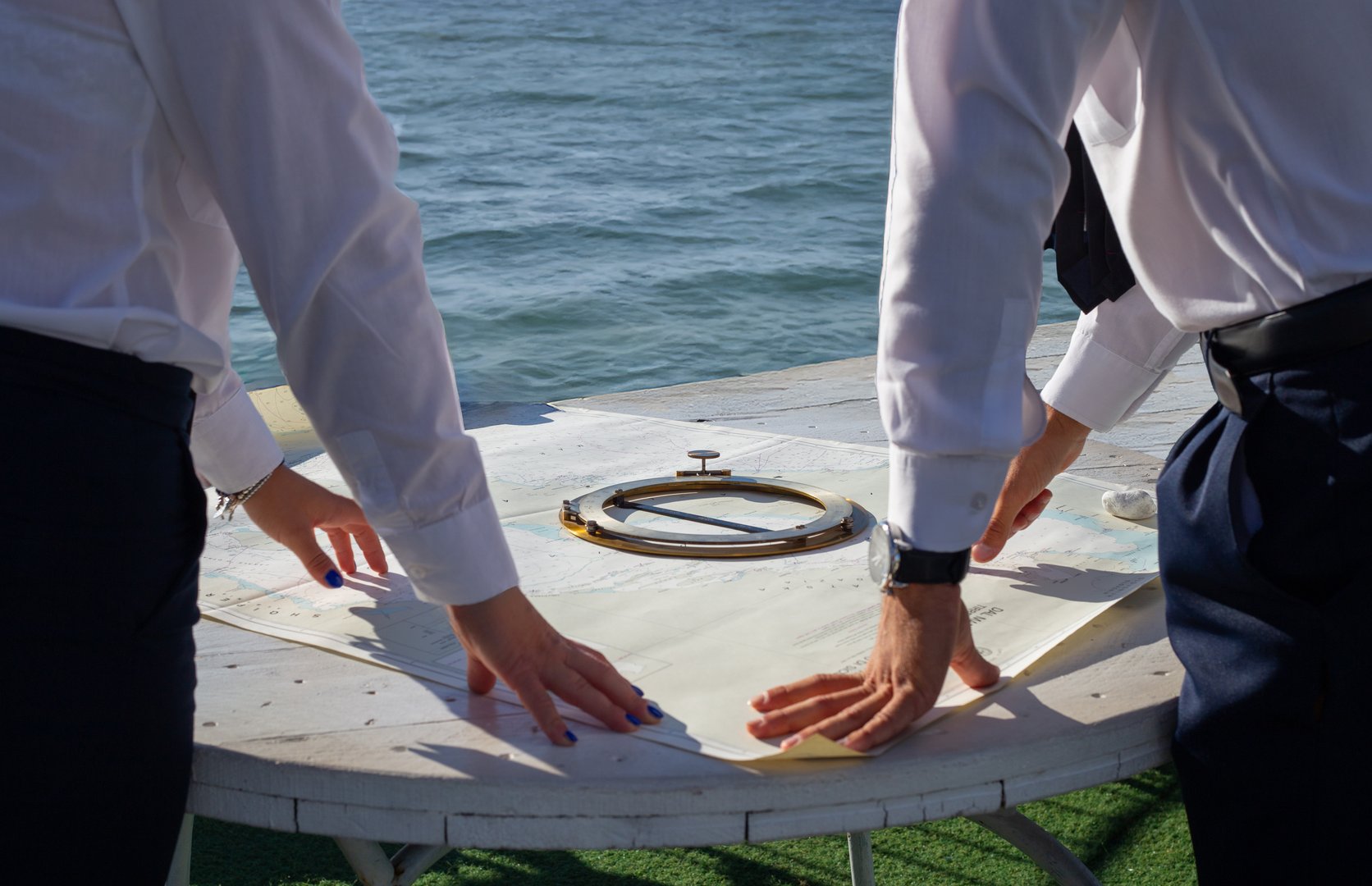 Two navigators in white shirts examine a nautical map on a white table, with a compass in the foreground and the sea in the background
