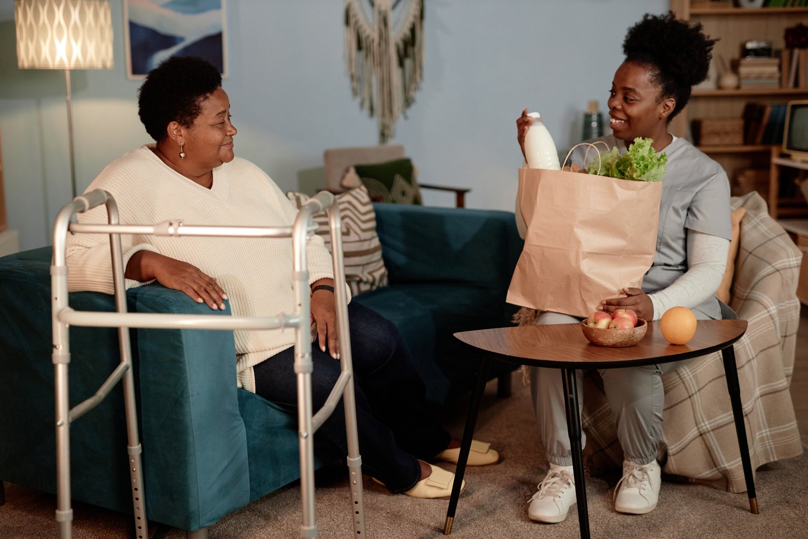 Full length shot of accommodating Black female social worker delivering groceries to senior womans house helping elderly people with physical disabilities, assisted living concept