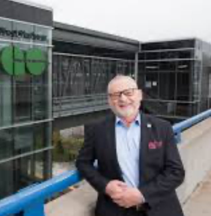 Man in a suit standing outdoors in front of a modern building with glass facade and a green logo.