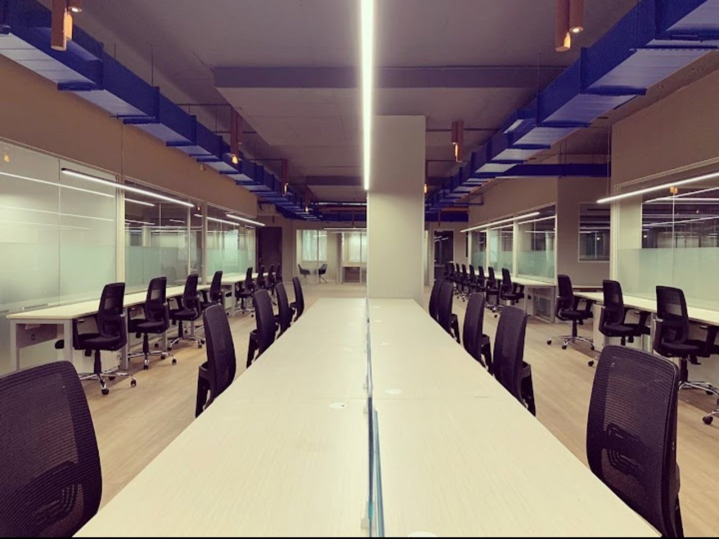 Modern office space with long tables, black chairs, and blue ceiling accents, surrounded by glass walls and partitions.