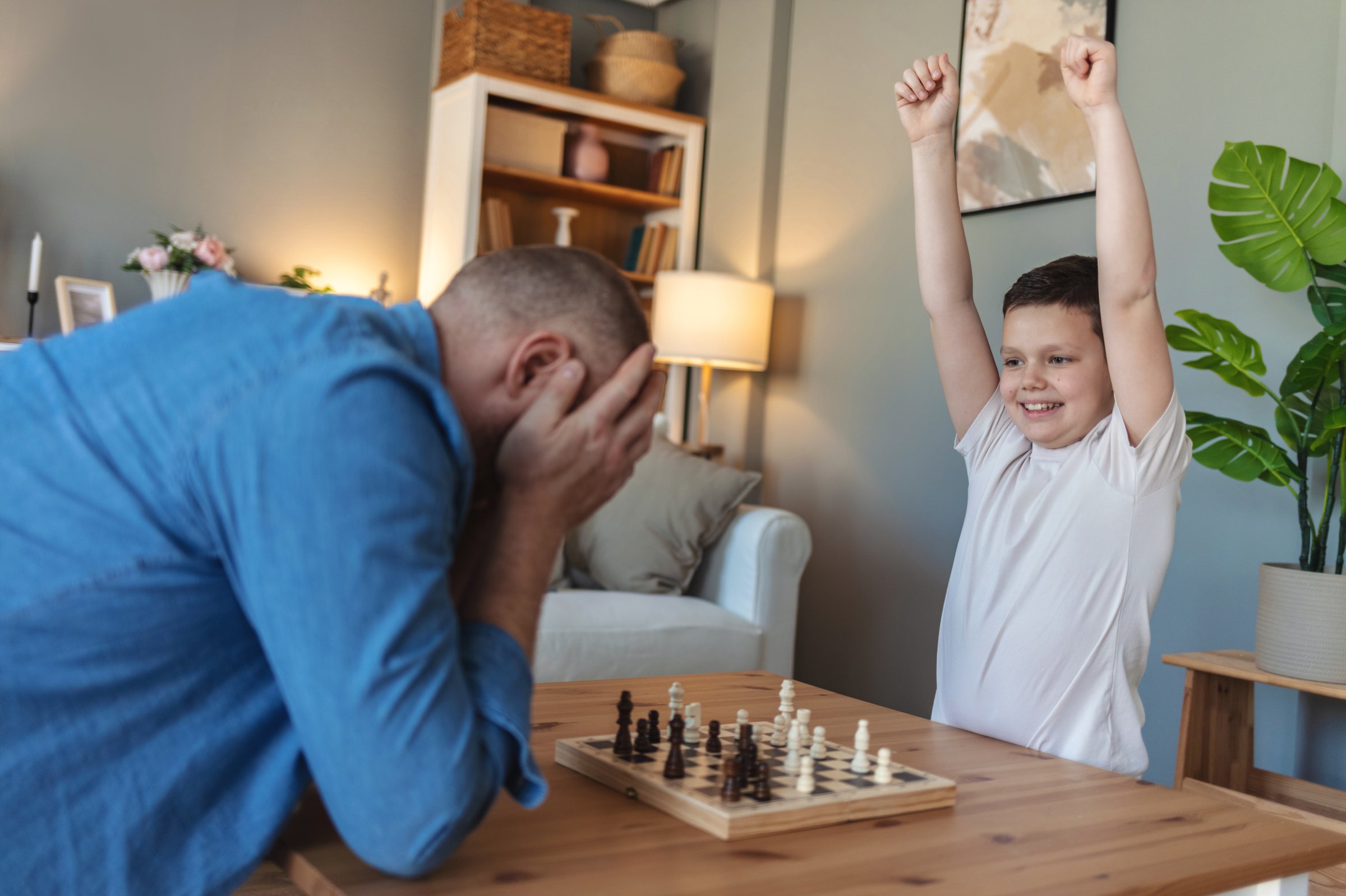 A delighted son raises his arms in celebration after winning a game of chess at home. His face lights up with joy and pride as he revels in his success, while the peaceful home setting adds to the warmth of this special moment of achievement.