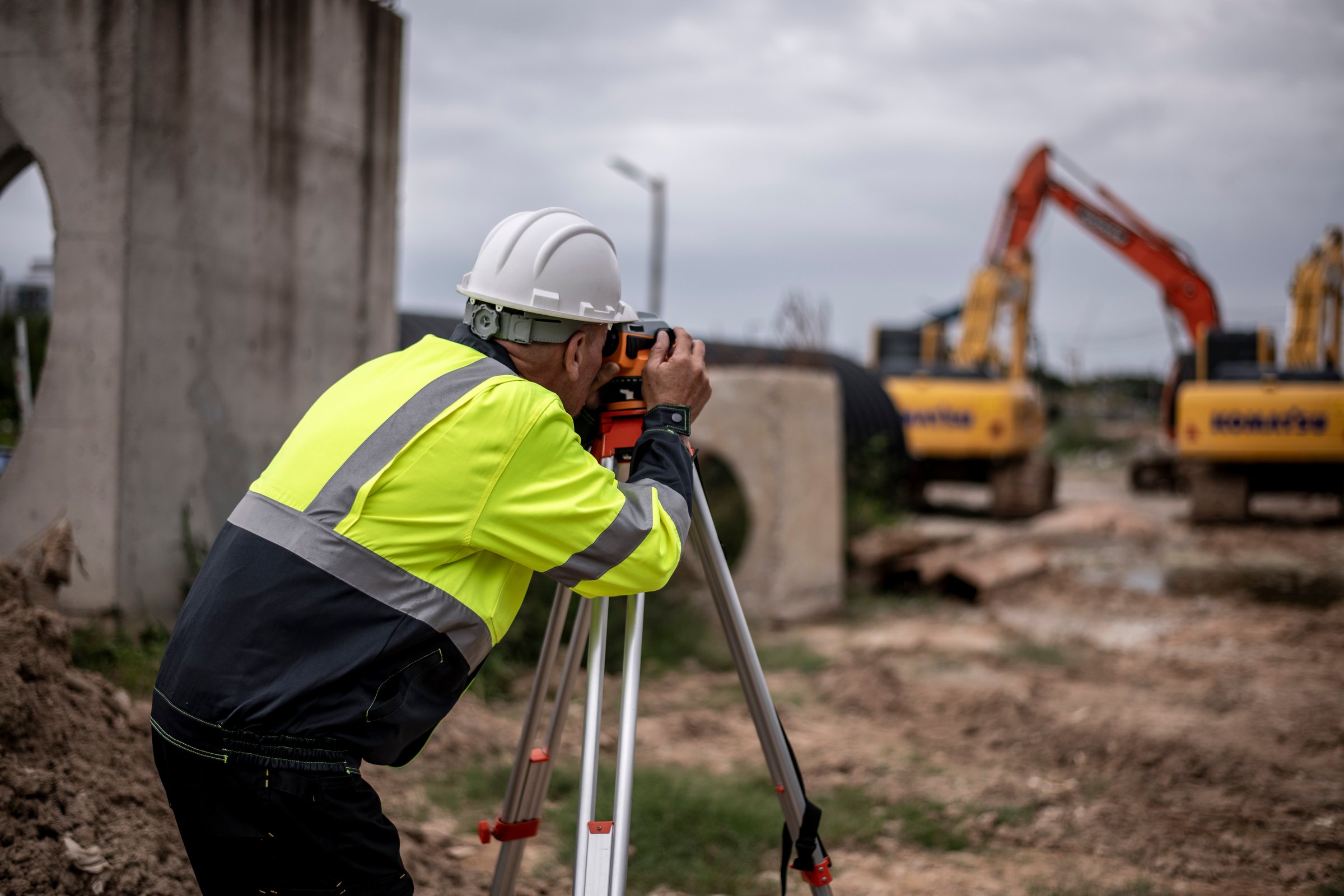 Construction workers wearing safety helmets and at a construction site. One worker is using a theodolite (or surveying equipment) on a tripod to take measurements, while the other worker holds a blueprint or set of construction plans.