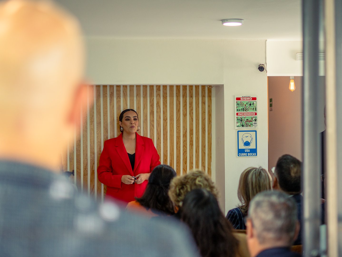 A woman in a red blazer speaks to a seated audience in a room with a wood-paneled wall and safety signs.