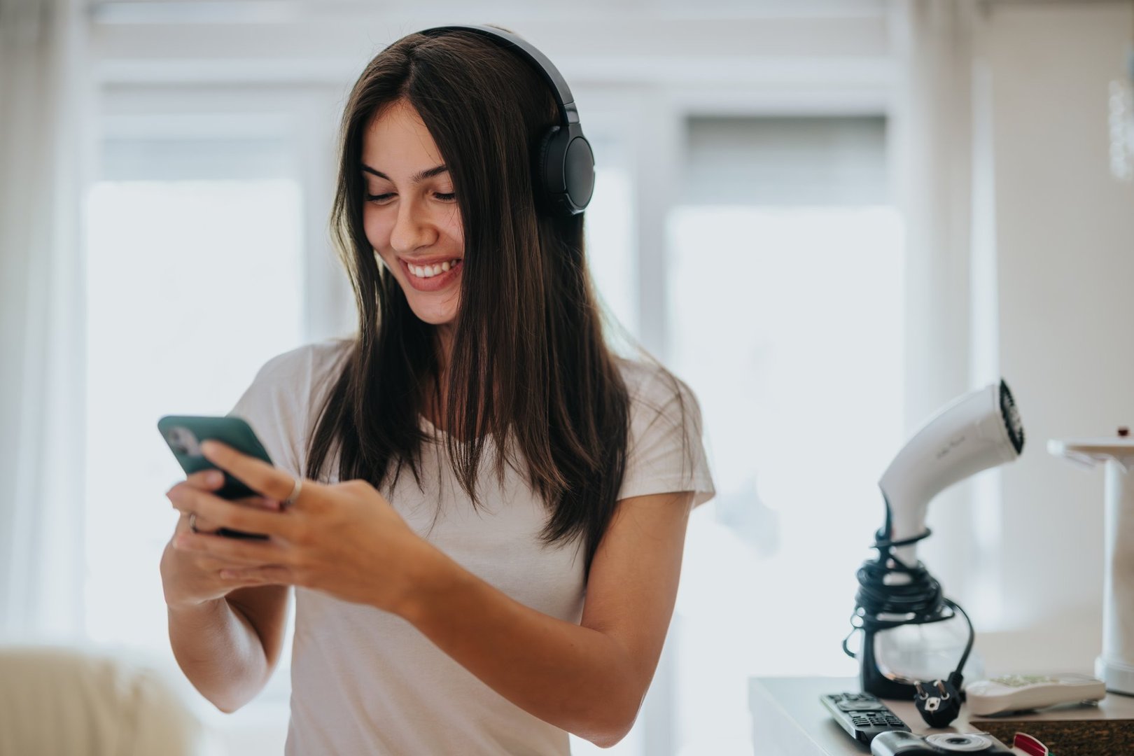 A cheerful young woman wearing headphones smiles as she uses her smartphone in a bright room. Her casual attire and joyful expression convey a relaxed and happy atmosphere.