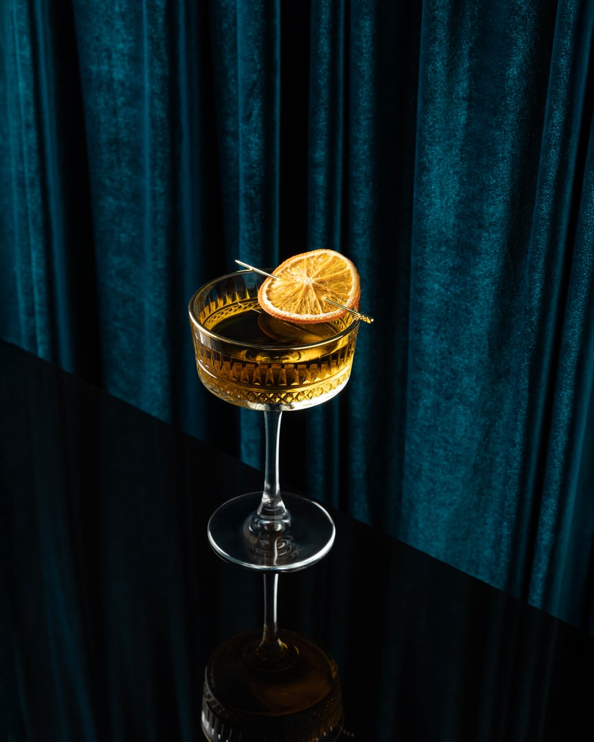 A crystal coupe glass with a golden cocktail garnished with a dried citrus slice, styled against rich teal velvet curtains and a glossy black reflective surface. Sophisticated, moody, and luxurious.
