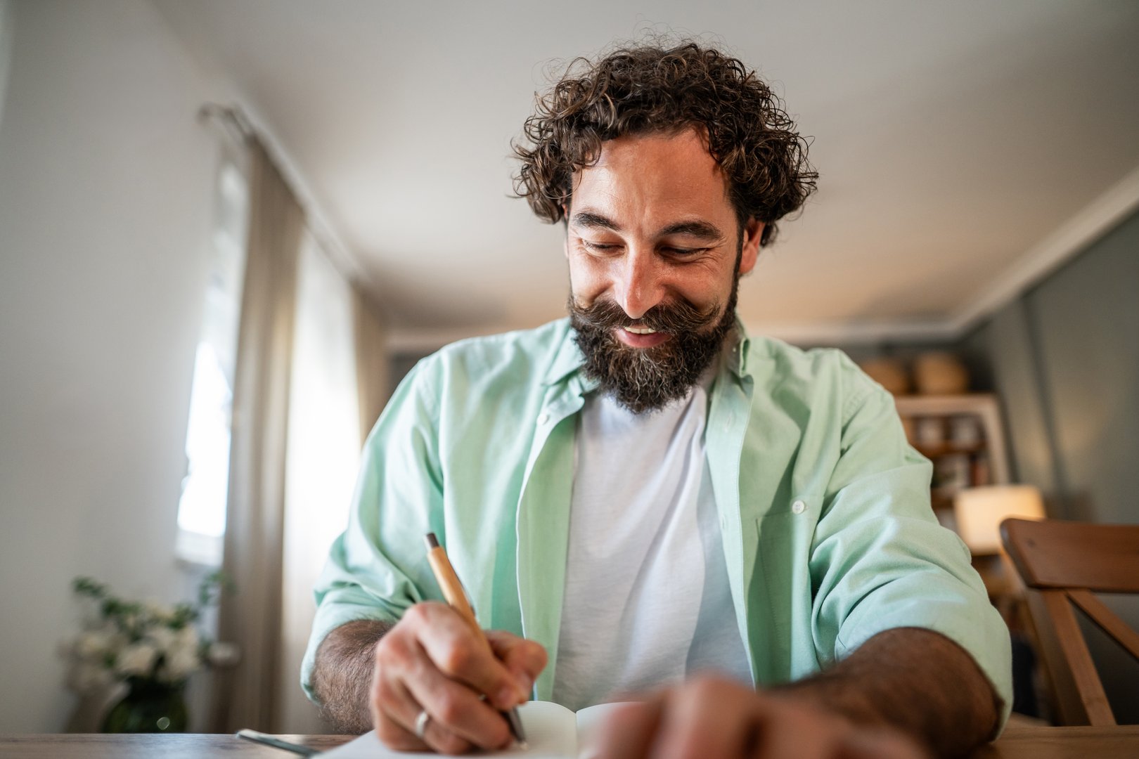 Cheerful adult man with a beard and mustache concentrating and smiling while writing thoughts and ideas in a personal journal notebook, enjoying a creative moment indoors