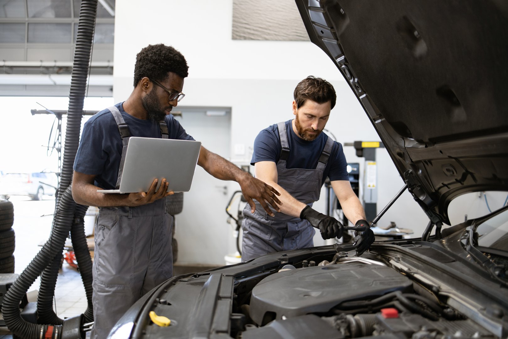Mechanics diagnosing car engine problem using laptop in modern garage setting. Technicians collaborating on vehicle repair in workshop environment, showcasing teamwork and technical skills.
