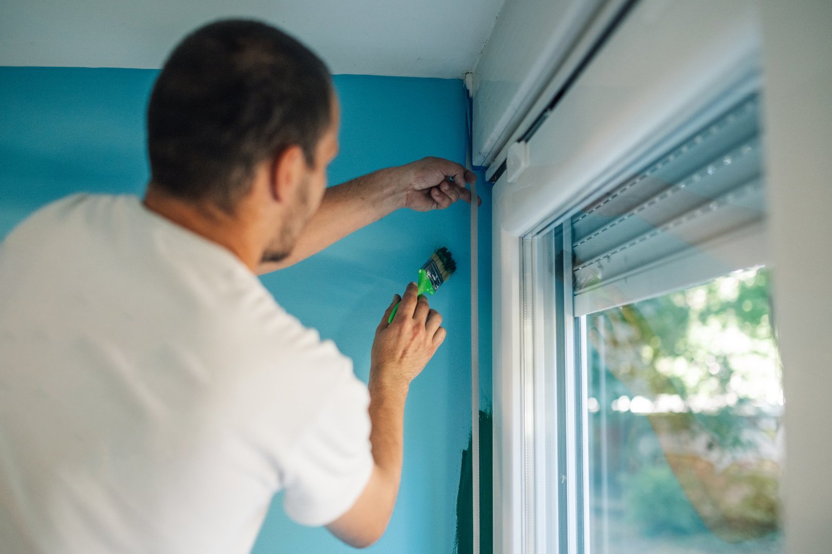 Painter applying masking tape around a window frame while skillfully painting a wall in a vibrant blue color, enhancing the room's decor