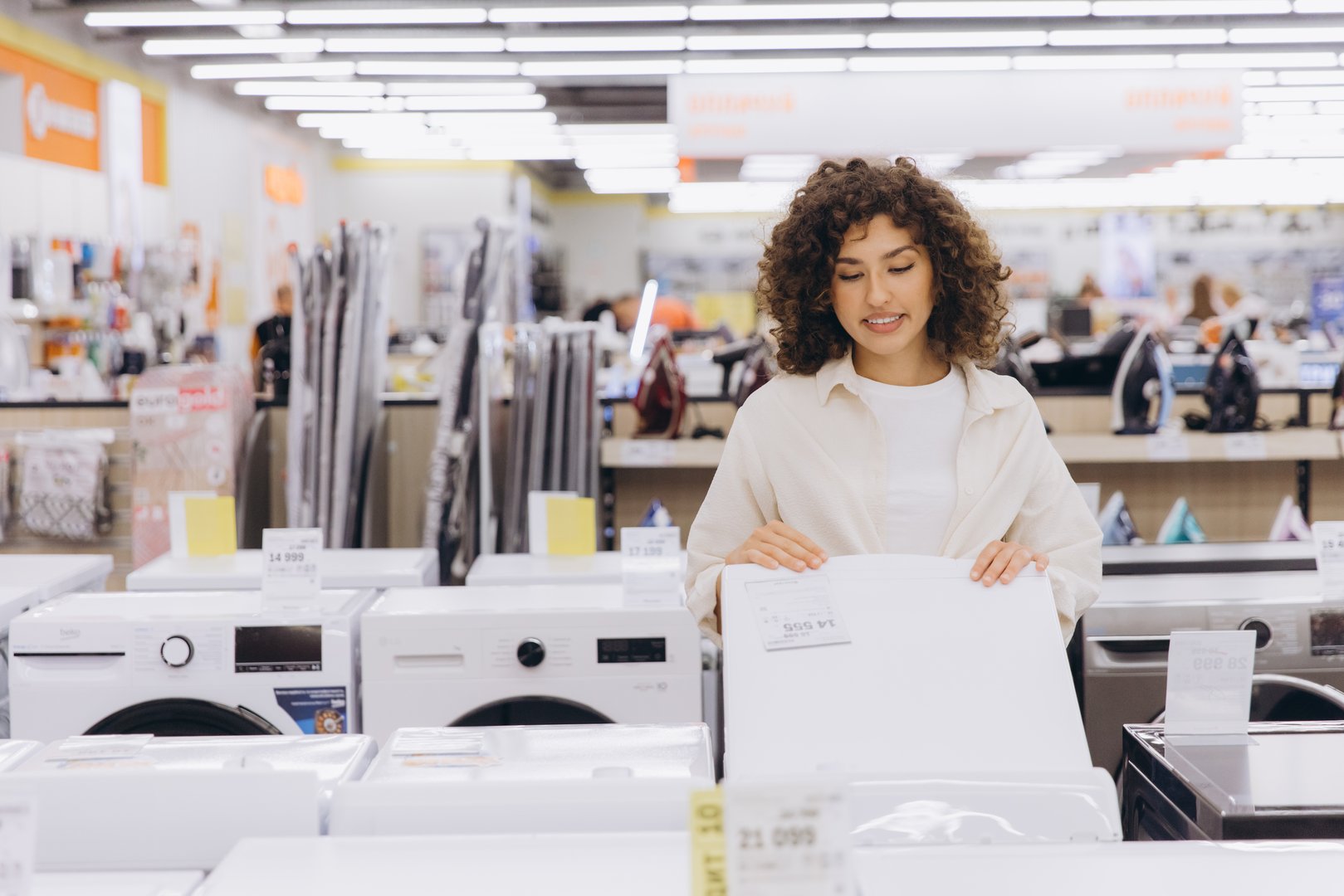 Young woman comparing washing machines, reading specifications and price tags in a home appliance store