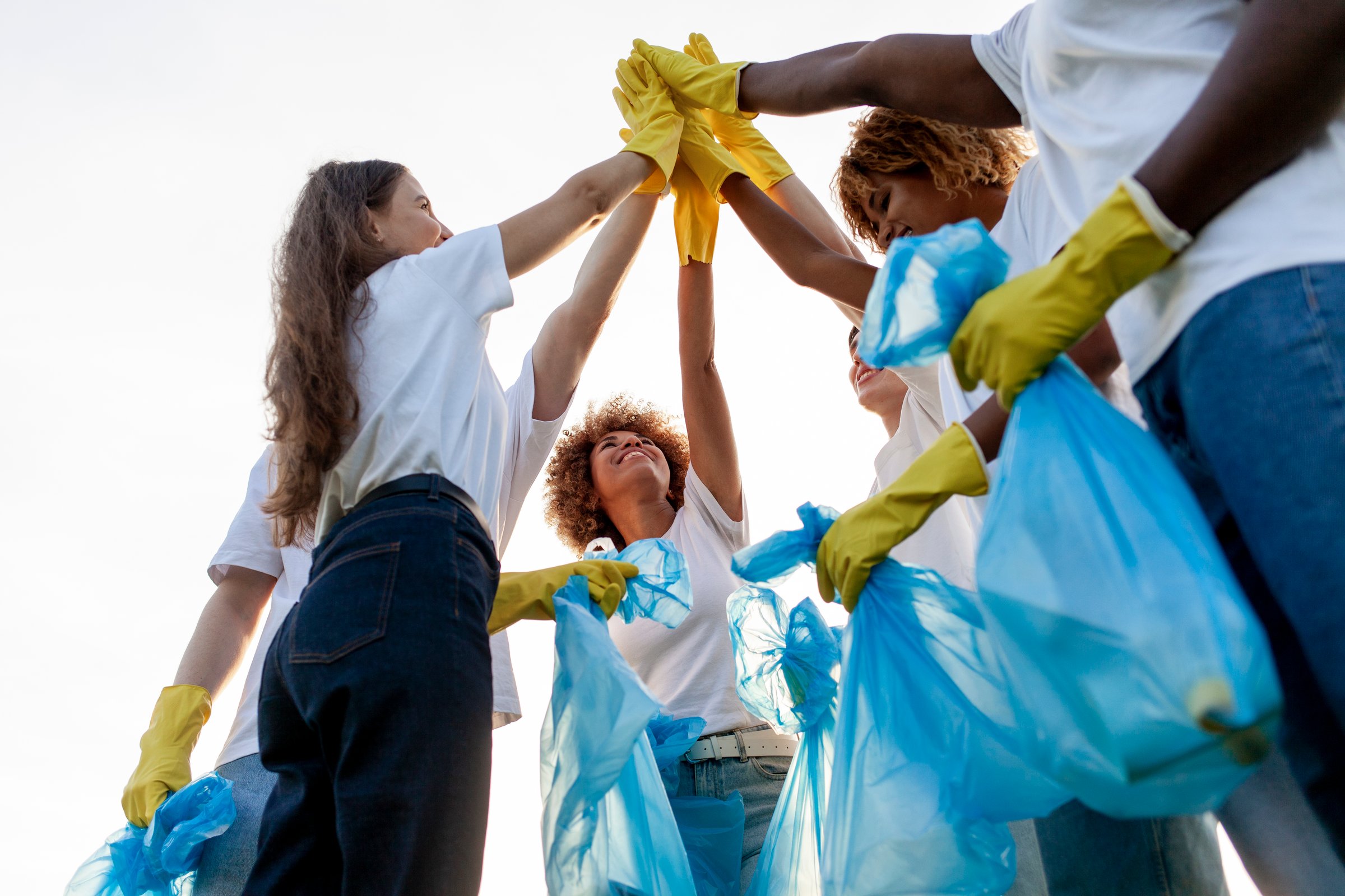group of multiracial people in gloves with garbage bags high-fiving and celebrating success together in the park, team of volunteers cleaning up trash and rejoicing in achievements